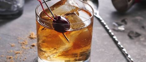 A glass of amber-colored beverage with ice cubes and a cherry on a metal straw.