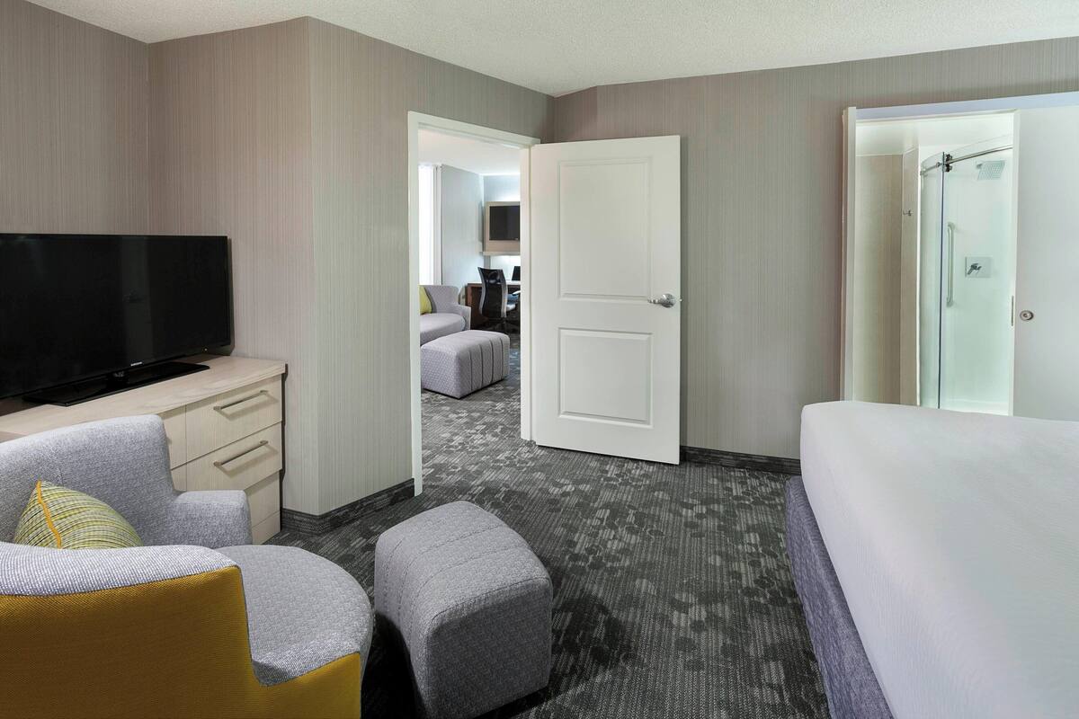 A hotel room with a flat-screen TV, a sofa, a small ottoman, a bed, a door leading to a bathroom, and a carpeted floor.