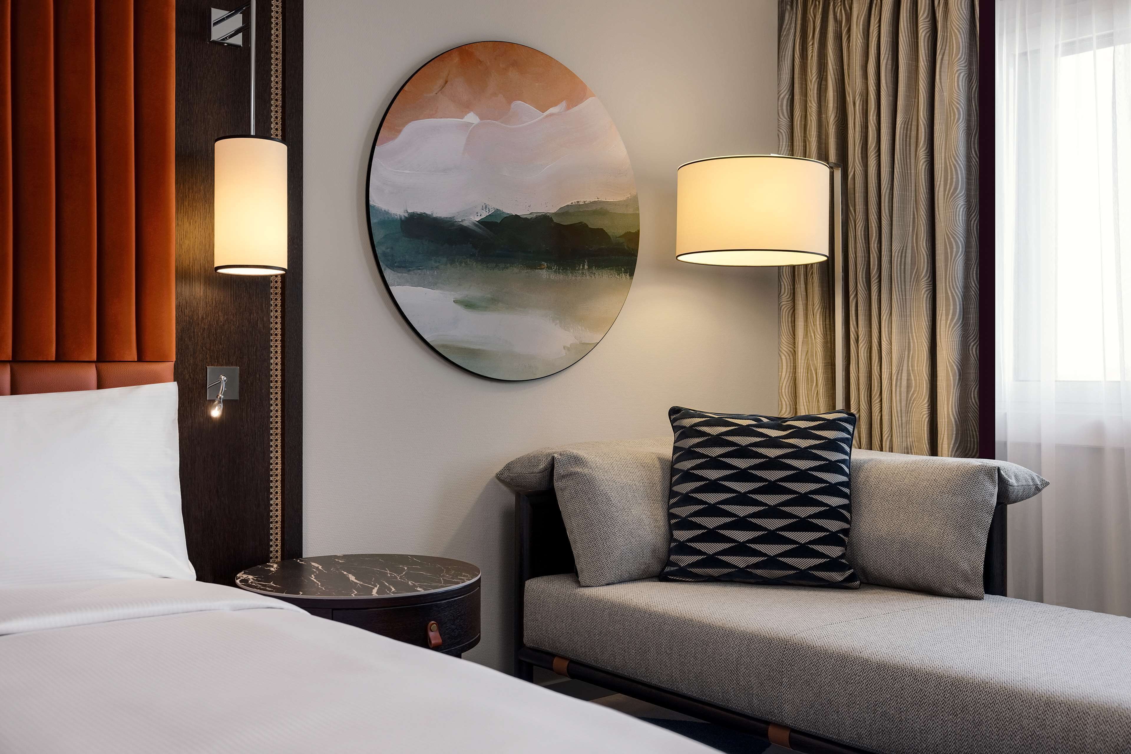 Habitación de hotel con cama, sofá, dos lámparas de pared, un cuadro redondo y una mesita de noche con lámpara.
