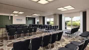 Uma sala de conferências com fileiras de cadeiras pretas, uma tela grande exibindo 'Hilton Vienna Waterfront' e amplas janelas com vista para o verde.