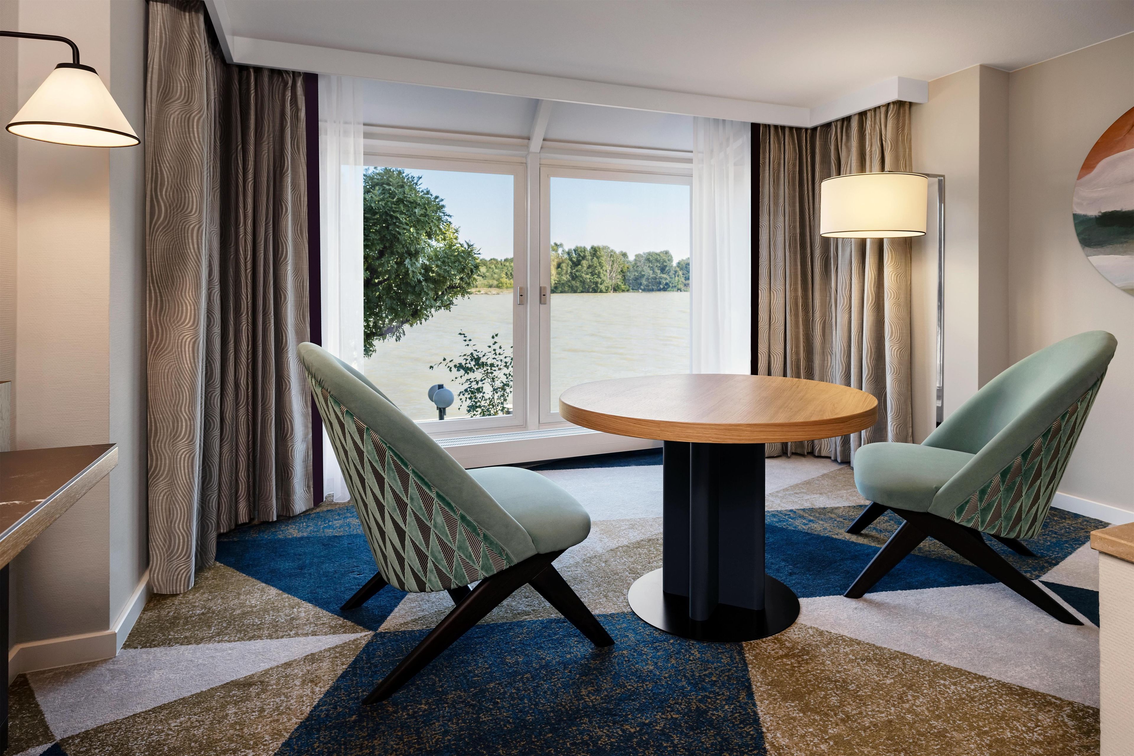 Ein Hotelzimmer mit einem runden Tisch, zwei Stühlen und Blick auf ein Gewässer, dazu ein großes Fenster.