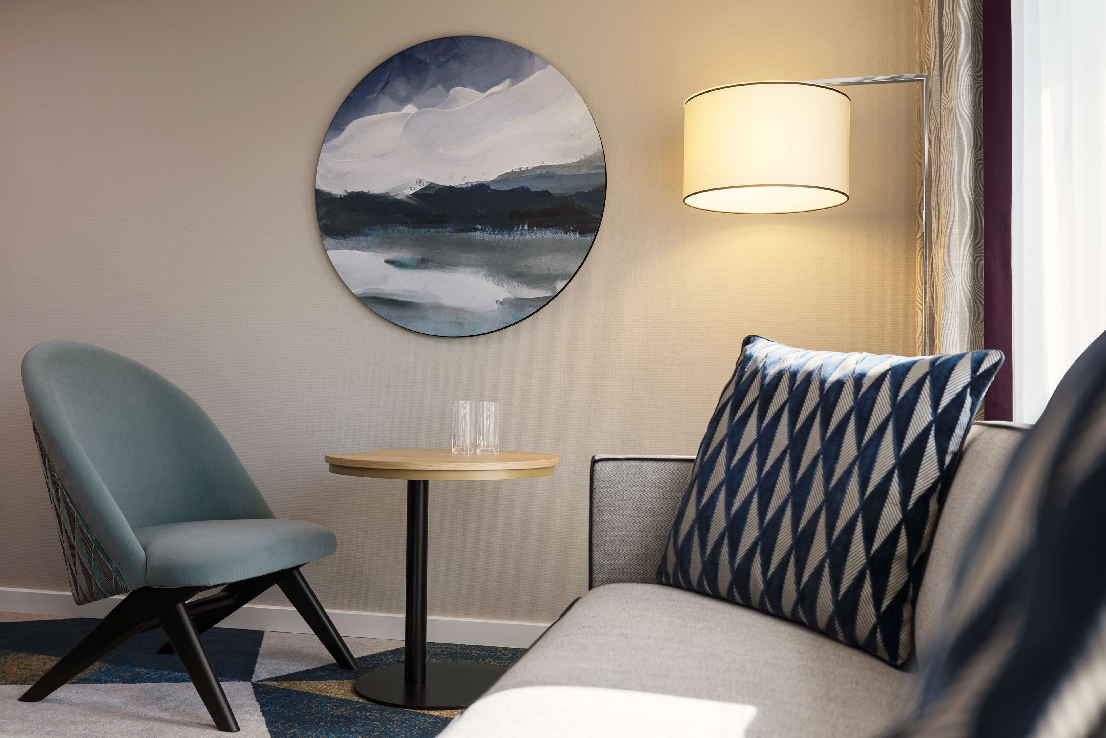Una habitación de hotel con un cuadro redondo de un paisaje, una mesa redonda con un vaso, un sofá con cojines estampados y una lámpara de pie.