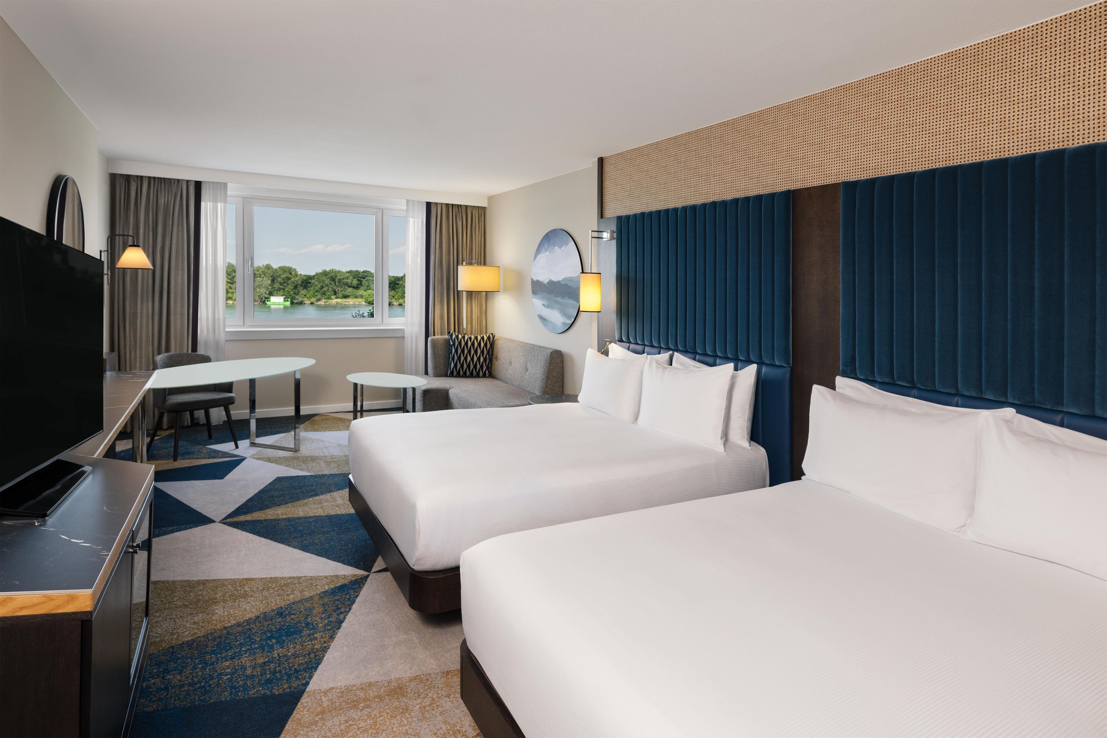 A hotel room with two beds, a desk, a TV, and a window with a view of greenery.