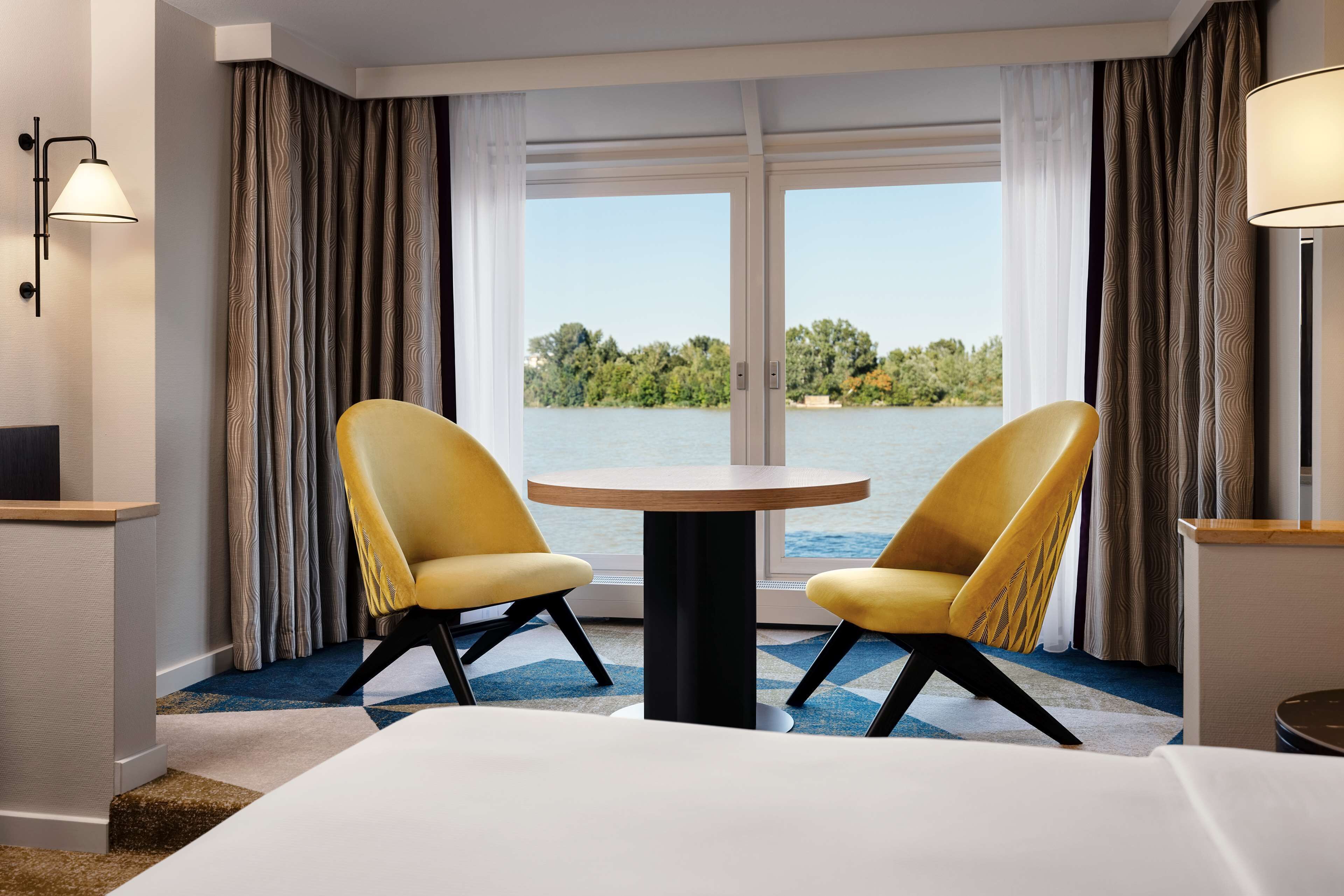 Ein Hotelzimmer mit großem Fenster, zwei gelben Stühlen, einem runden Tisch und einem Bett.