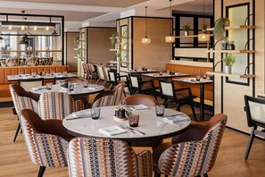 A restaurant interior with round tables, patterned chairs, and a bar area with shelves and plants. - Hilton Vienna Waterfront (Vienna)