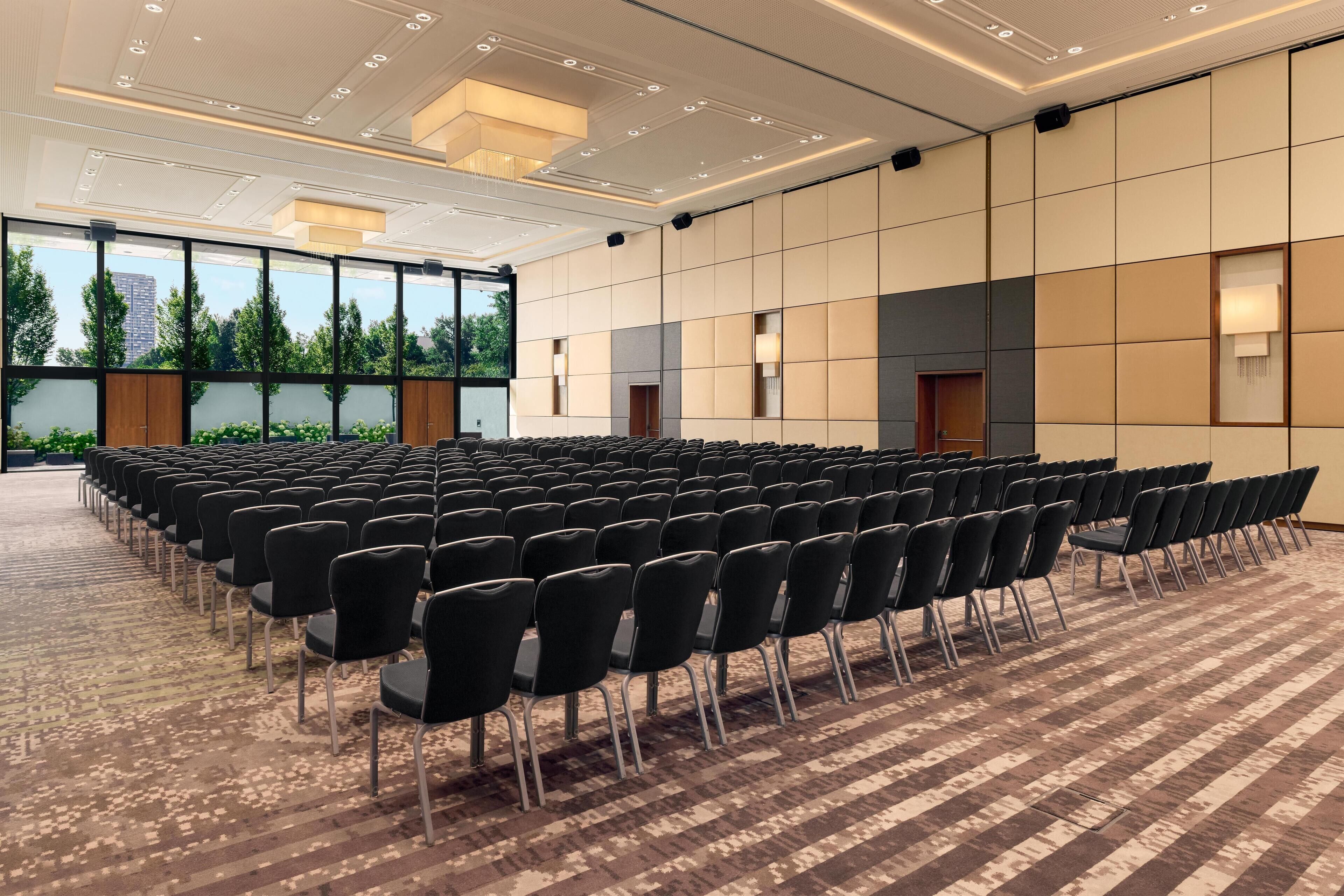 Un salón de conferencias con filas de sillas, ventanas amplias y una alfombra estampada.