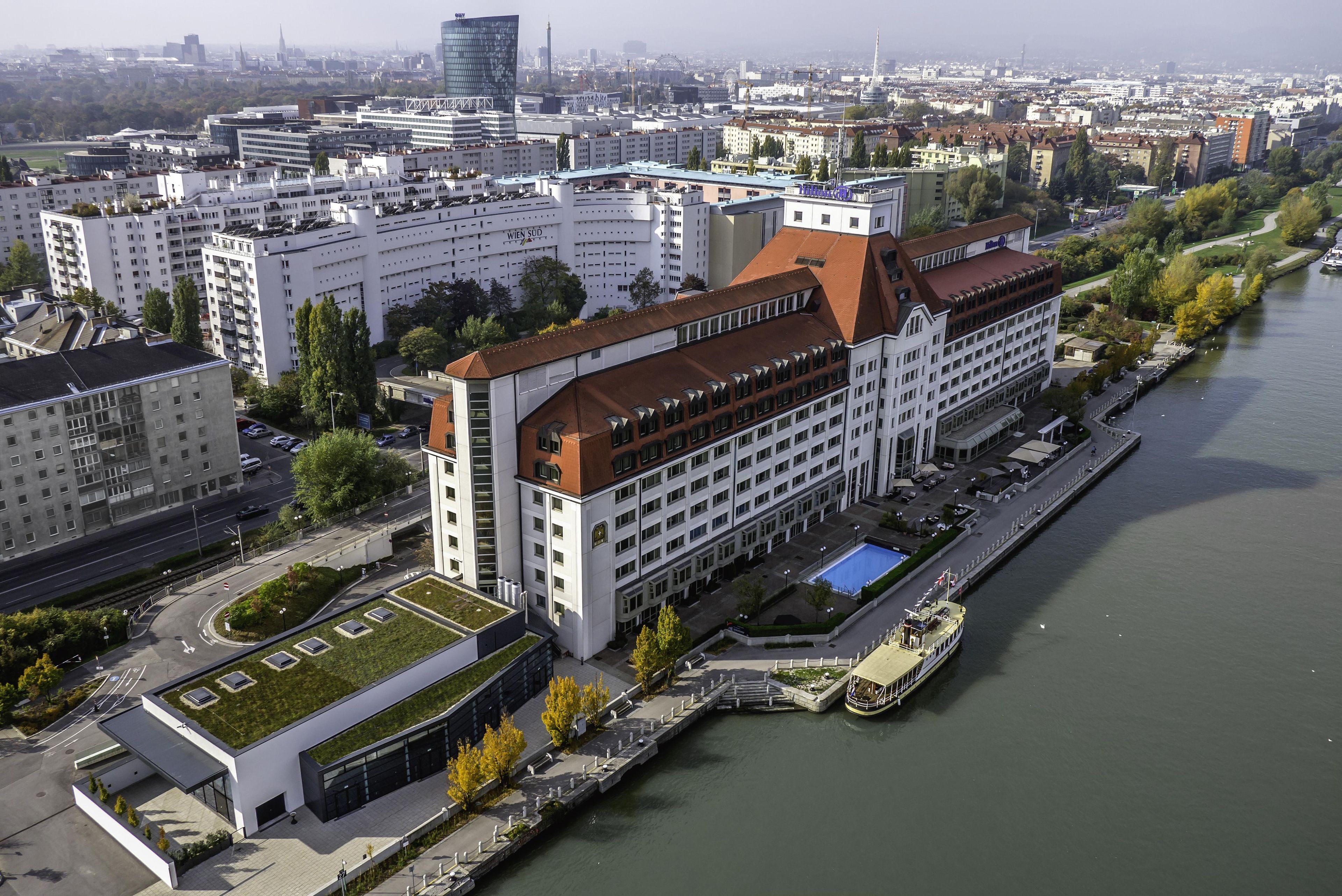 A hotel with a modern design, located beside a river. It features a prominent building with a red roof and a swimming pool.