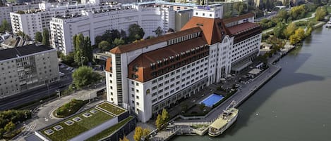 A hotel with a modern design, located beside a river. It features a prominent building with a red roof and a swimming pool.