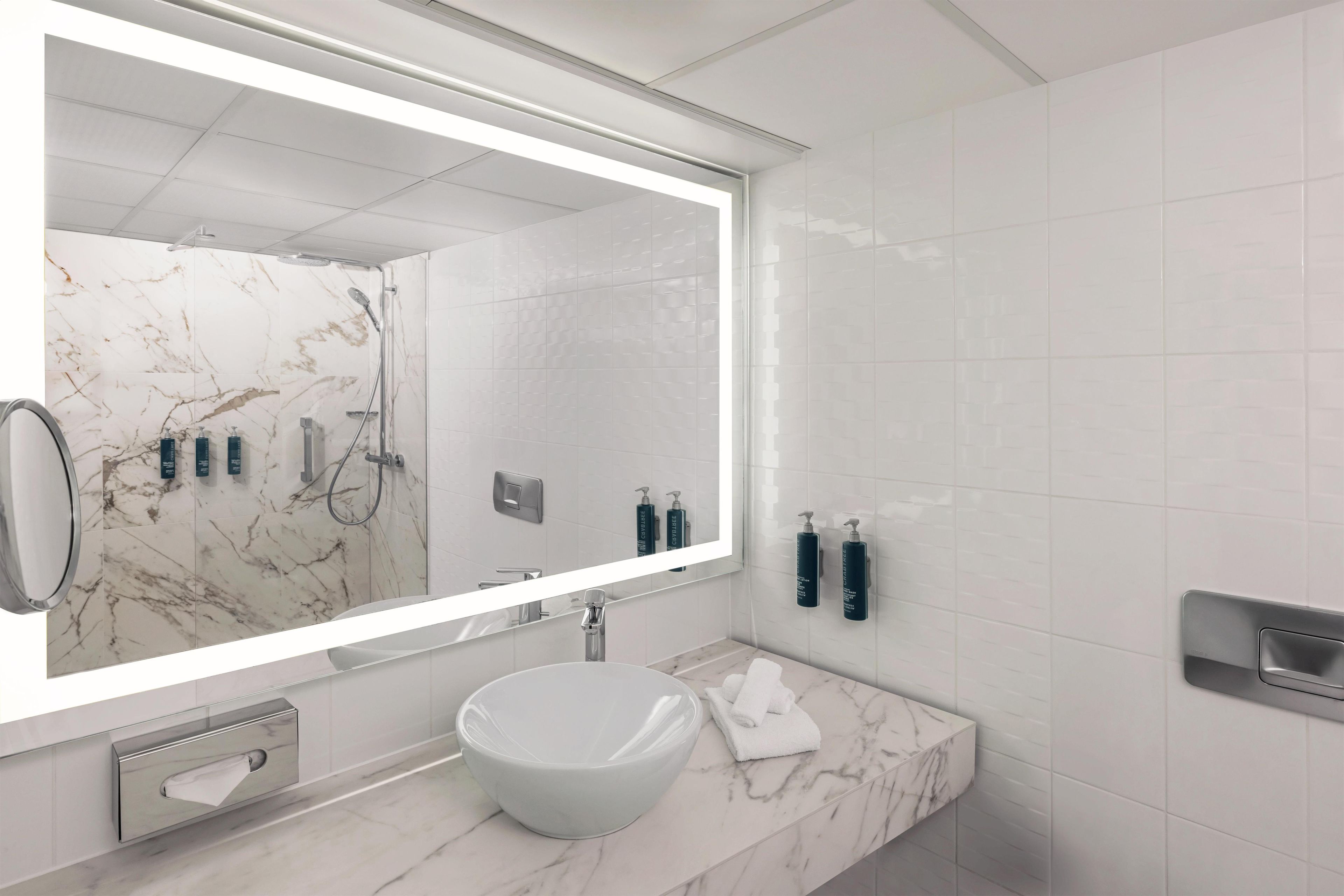 Une salle de bain moderne avec un grand miroir, un lavabo blanc et des carreaux muraux qui imitent le marbre.