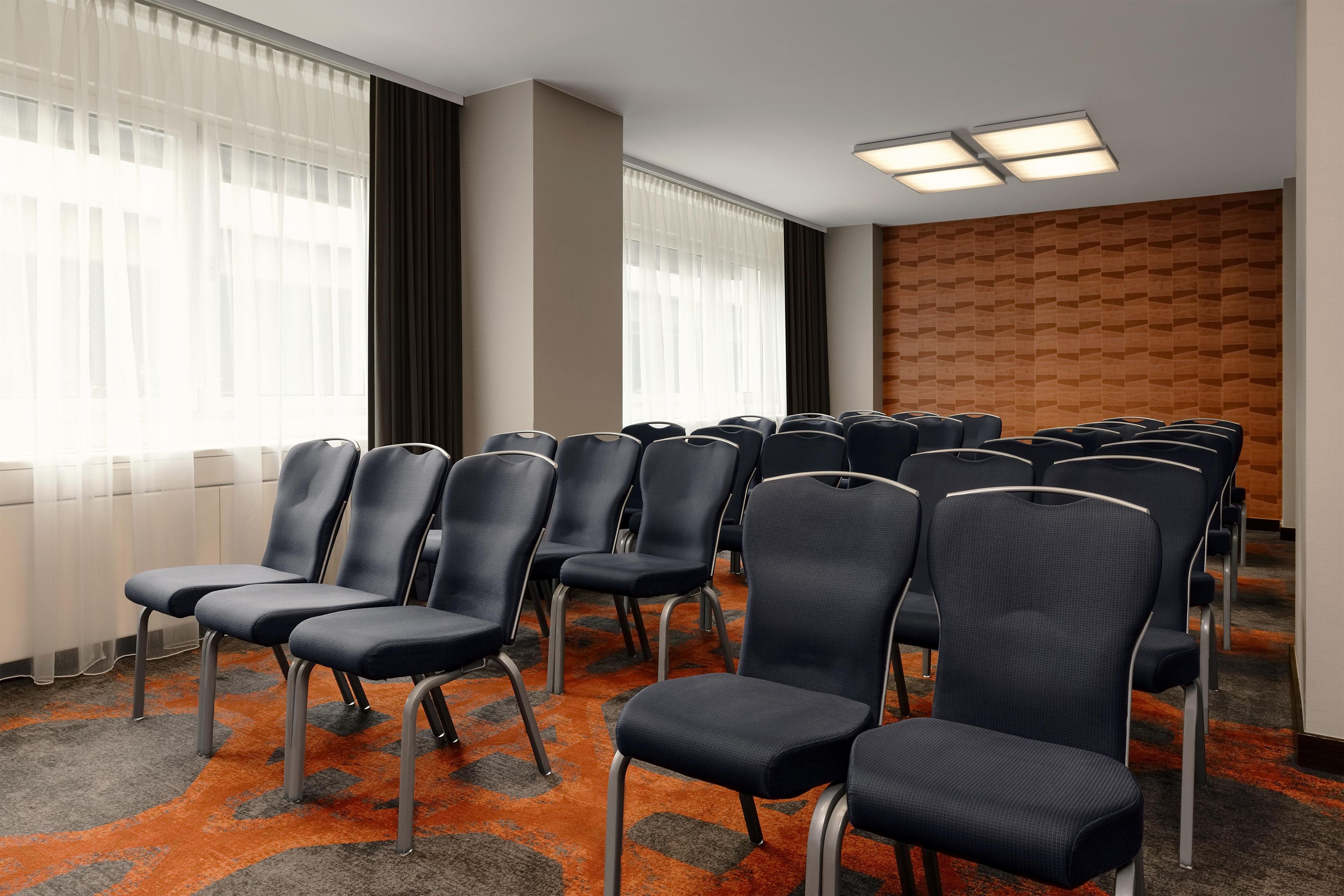 Sala de conferências com fileiras de cadeiras, amplas janelas com cortinas leves e carpete estampado.