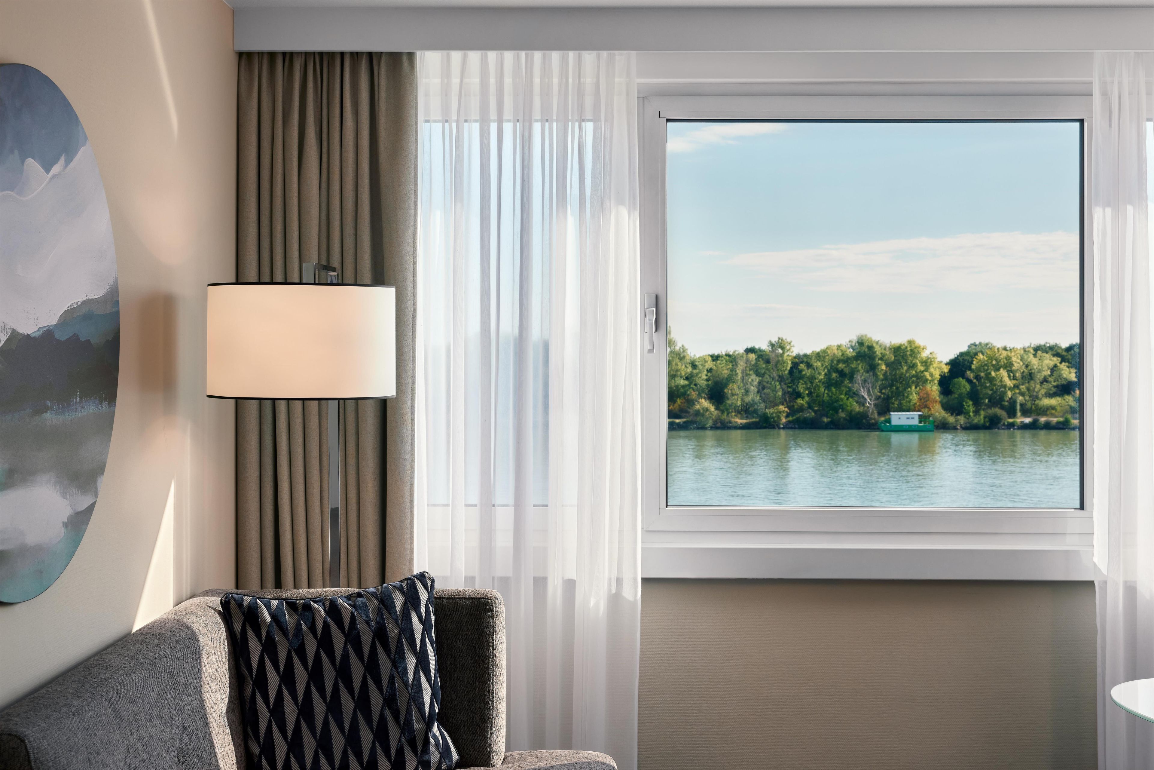 A hotel room with a view of a lake, a sofa, a lamp, and sheer curtains.