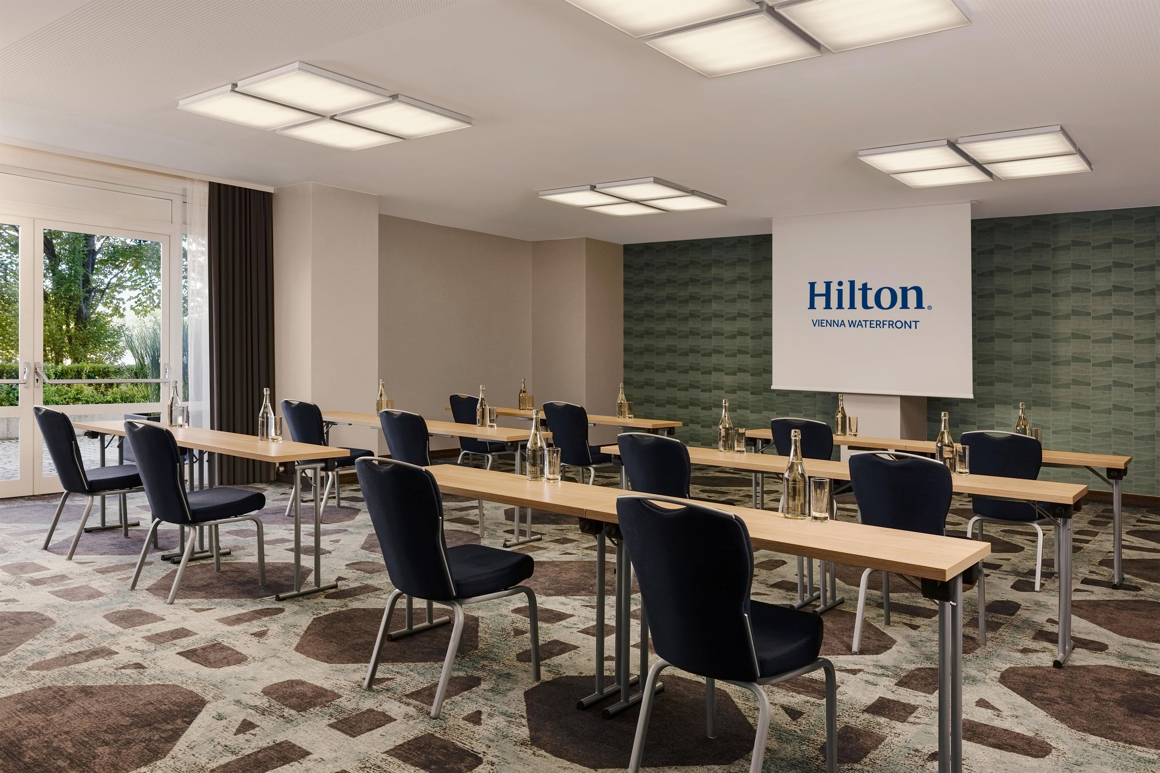A conference room with a long table, chairs, and a projector screen displaying 'Hilton Vienna Waterfront'.