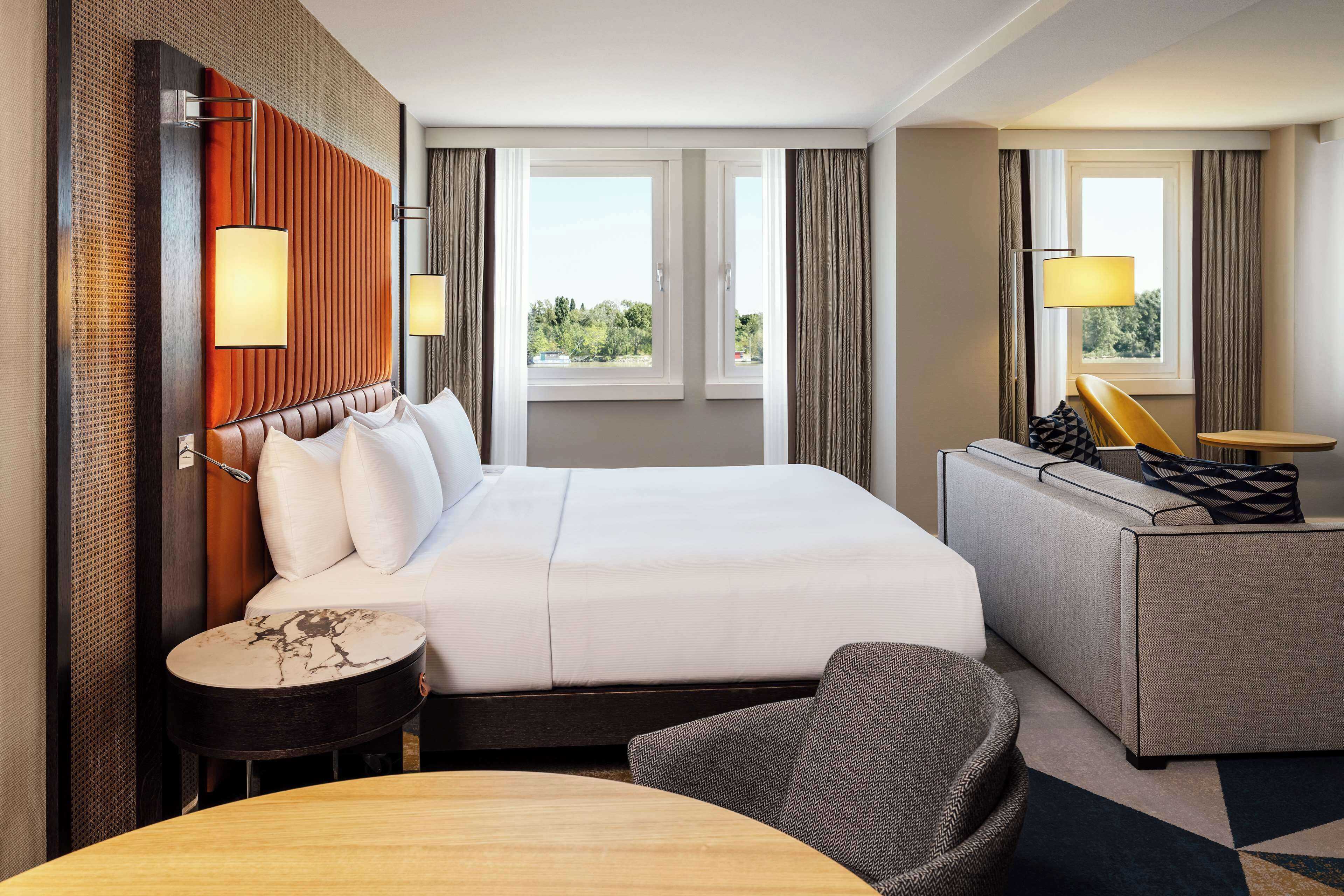 Ein Hotelzimmer mit einem großen Bett, einer Couch, einem kleinen runden Tisch und einem Sessel. Zwei Fenster mit Vorhängen und eine Wandlampe sind vorhanden.