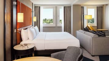 Ein Hotelzimmer mit einem großen Bett, einer Couch, einem kleinen runden Tisch und einem Sessel. Zwei Fenster mit Vorhängen und eine Wandlampe sind vorhanden.