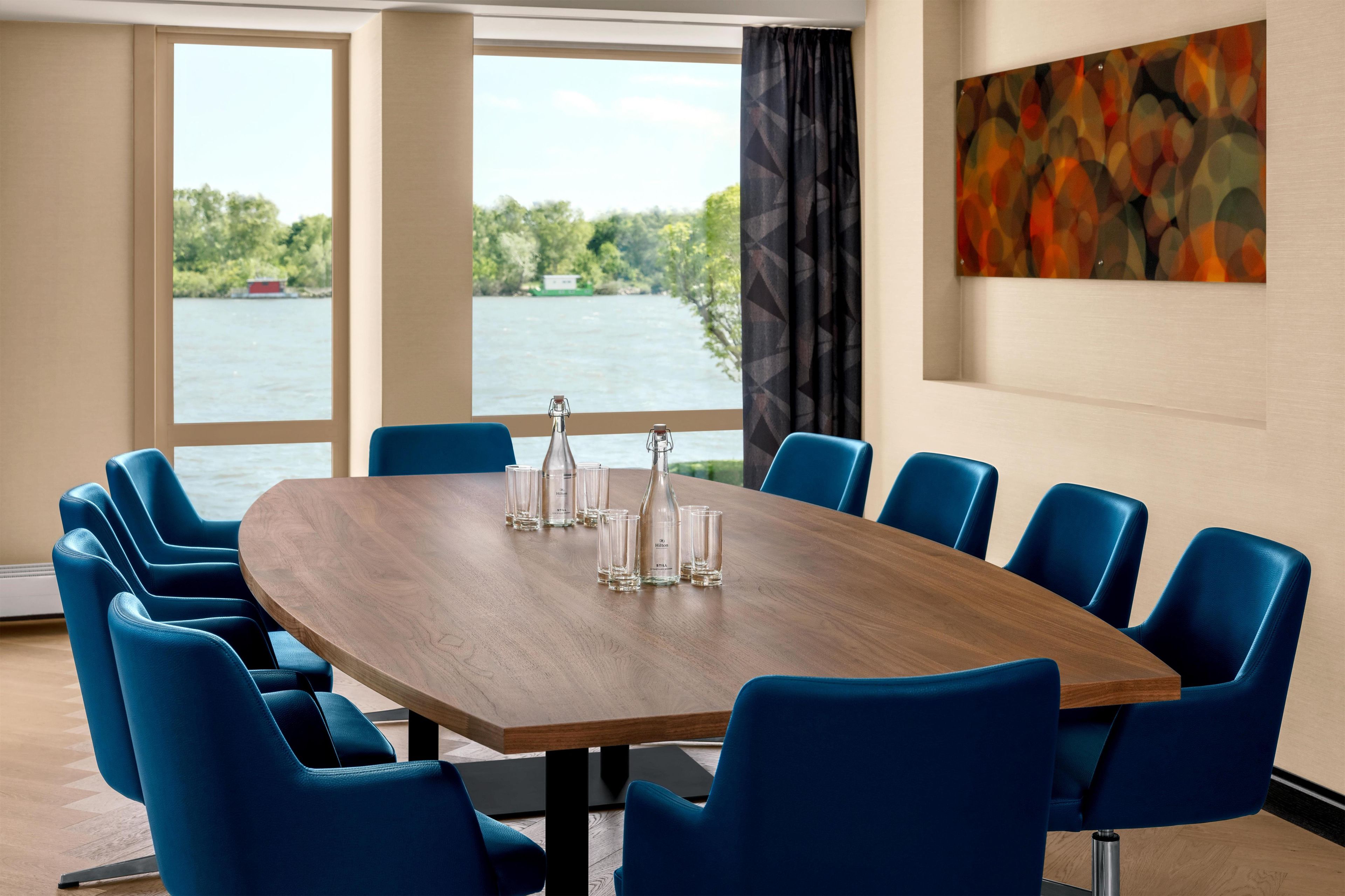 Un salón de conferencias con una gran mesa de madera, sillas azules y vistas a un lago a través de amplias ventanas.