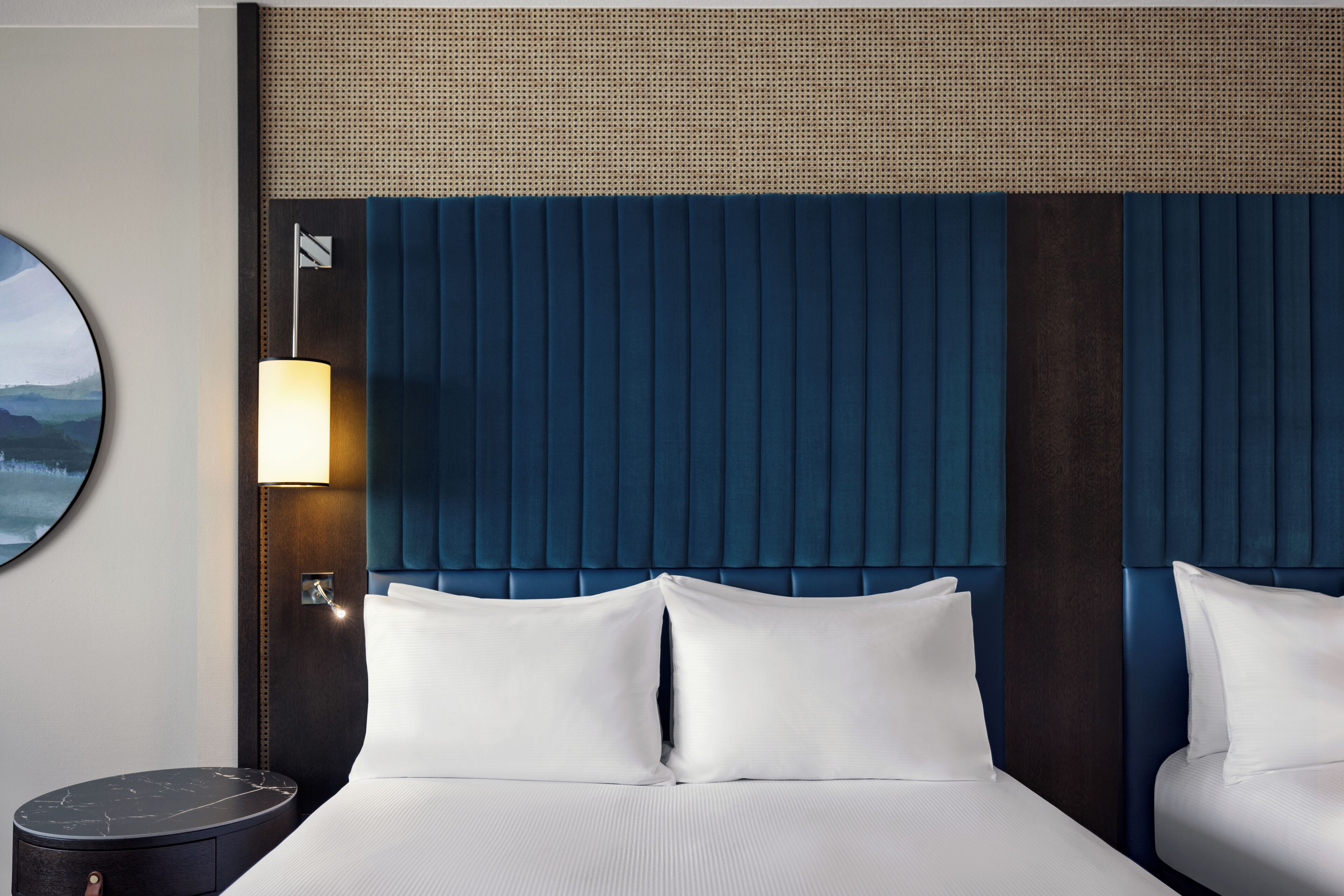 Quarto de hotel com uma cama grande, duas almofadas, uma mesa de cabeceira, um abajur e um quadro na parede.