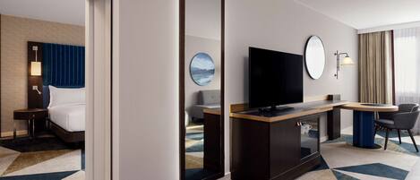 Habitación de hotel con cama, escritorio, televisor, espejo y vistas al exterior.