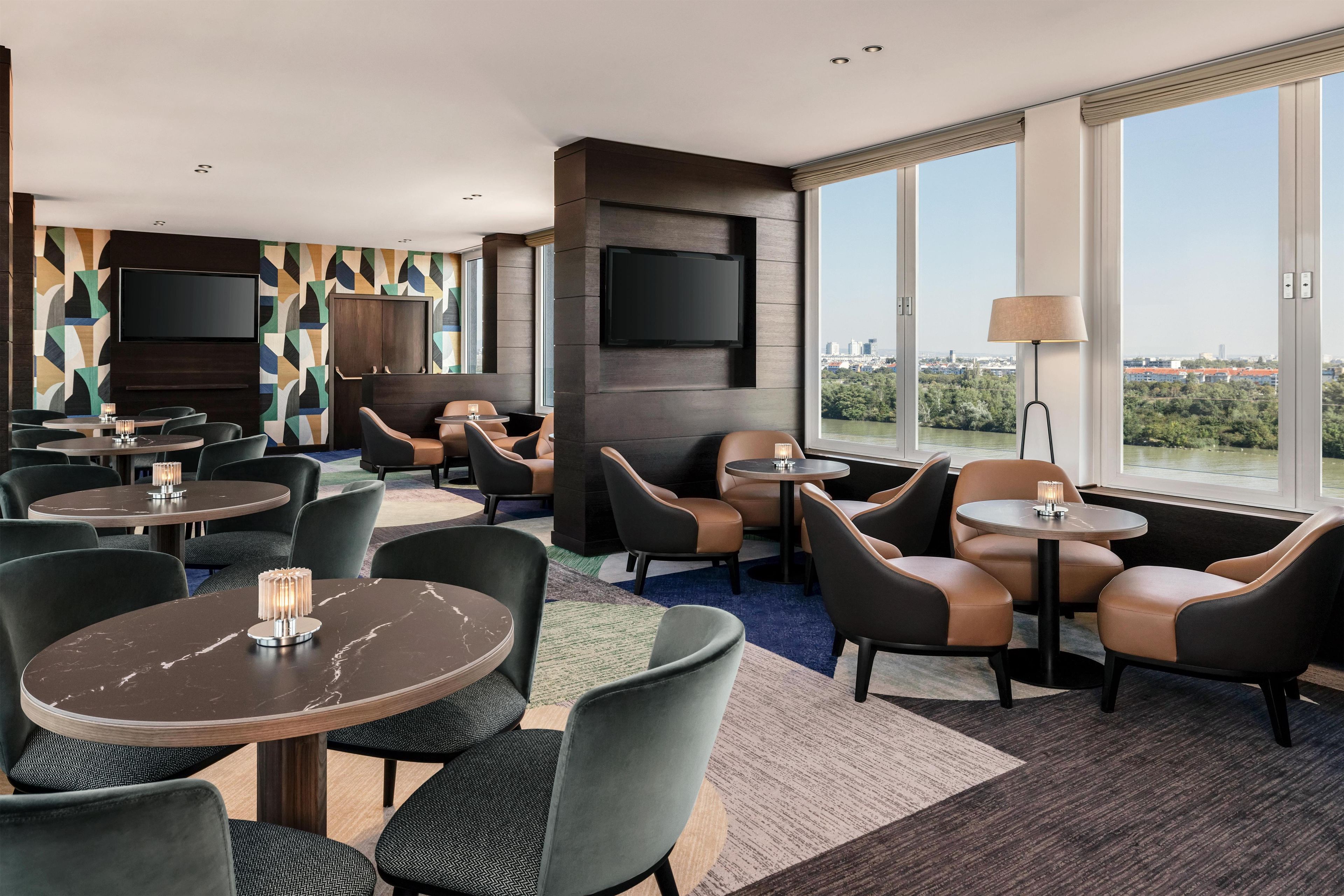 Un hall d’hôtel moderne avec des tables rondes, des fauteuils en cuir, un téléviseur à écran plat et une grande fenêtre donnant sur la ville.