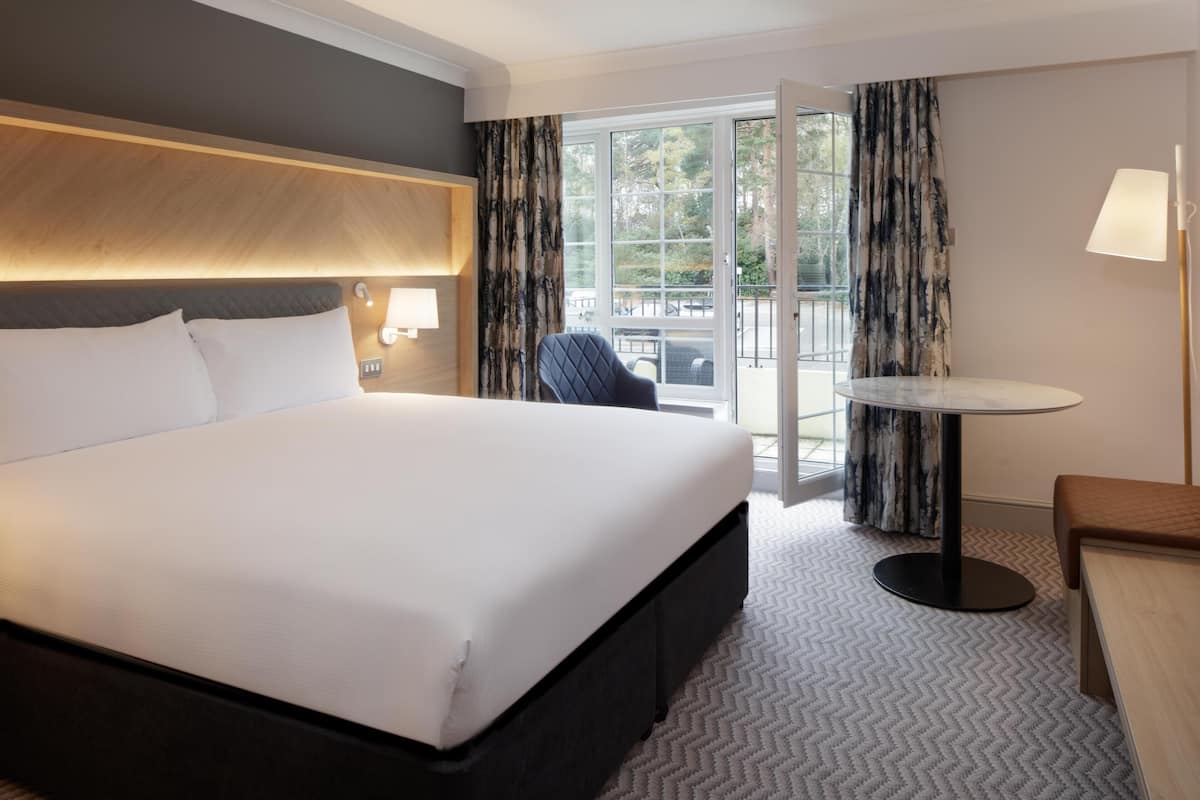 A hotel room with a large bed, a small table, a chair, a lamp, and a view of the outdoors through a sliding door.