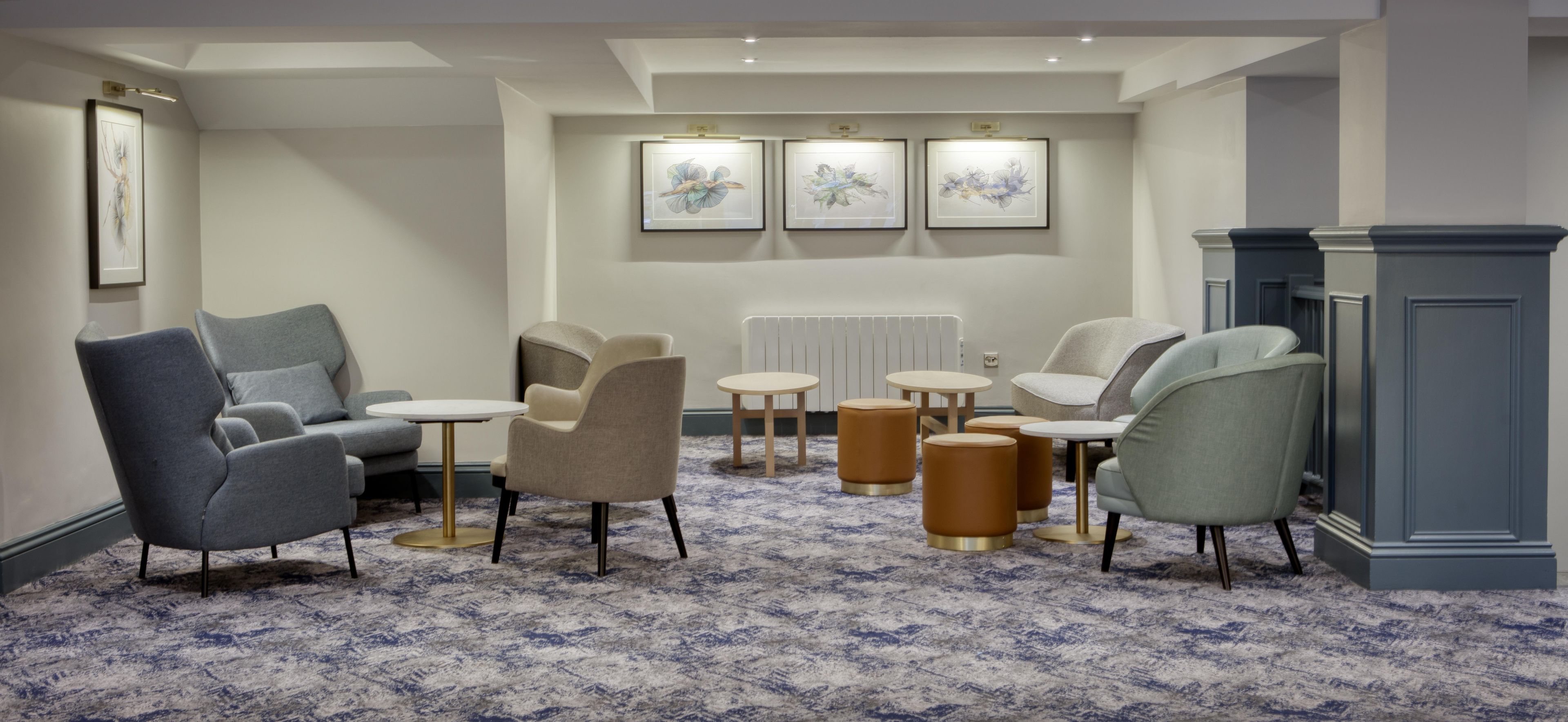 A modern lounge area with sofas, armchairs, and small tables, featuring a patterned carpet and framed artwork on the wall.