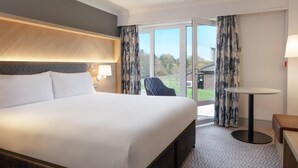 A hotel room with a large bed, a small table, a chair, and a view of the outdoors through a glass door. - Hilton Cobham (Cobham)