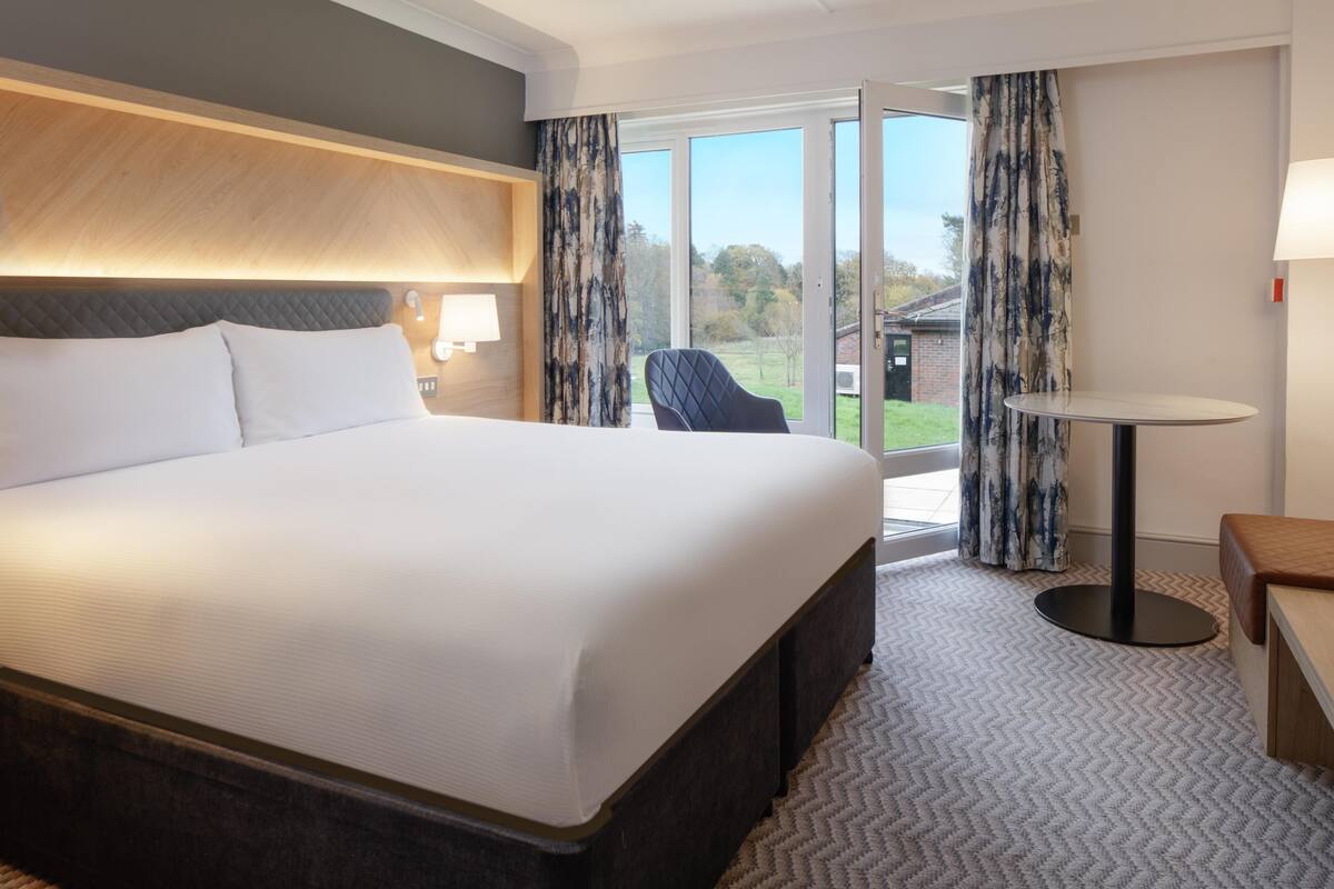A hotel room with a large bed, a small table, a chair, and a view of the outdoors through a glass door.
