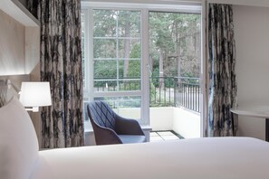 A hotel room with a bed, a chair, a table, and a balcony with a view of trees.