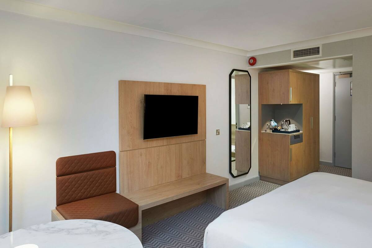 A hotel room with a bed, a seating area with a chair, a TV mounted on a wooden panel, a mirror, and a small shelf with items.