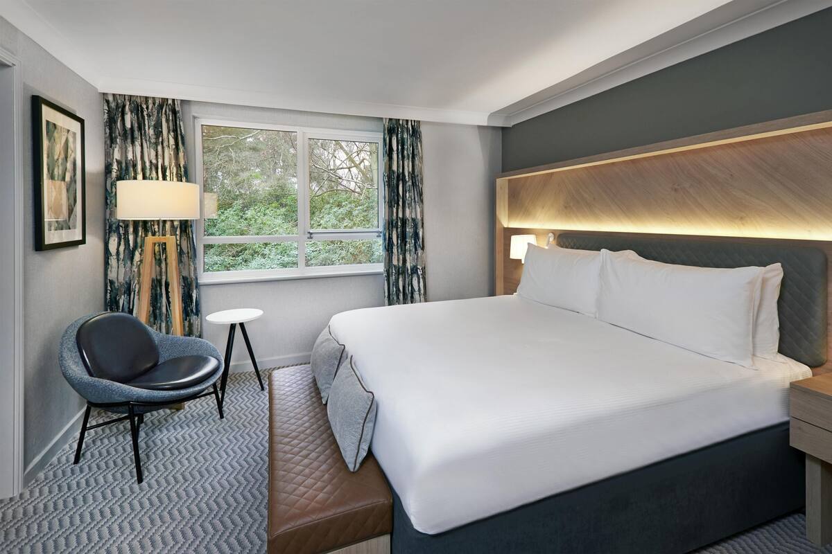 A hotel room with a large bed, a chair, a small table, a window with curtains, and a headboard with a light.