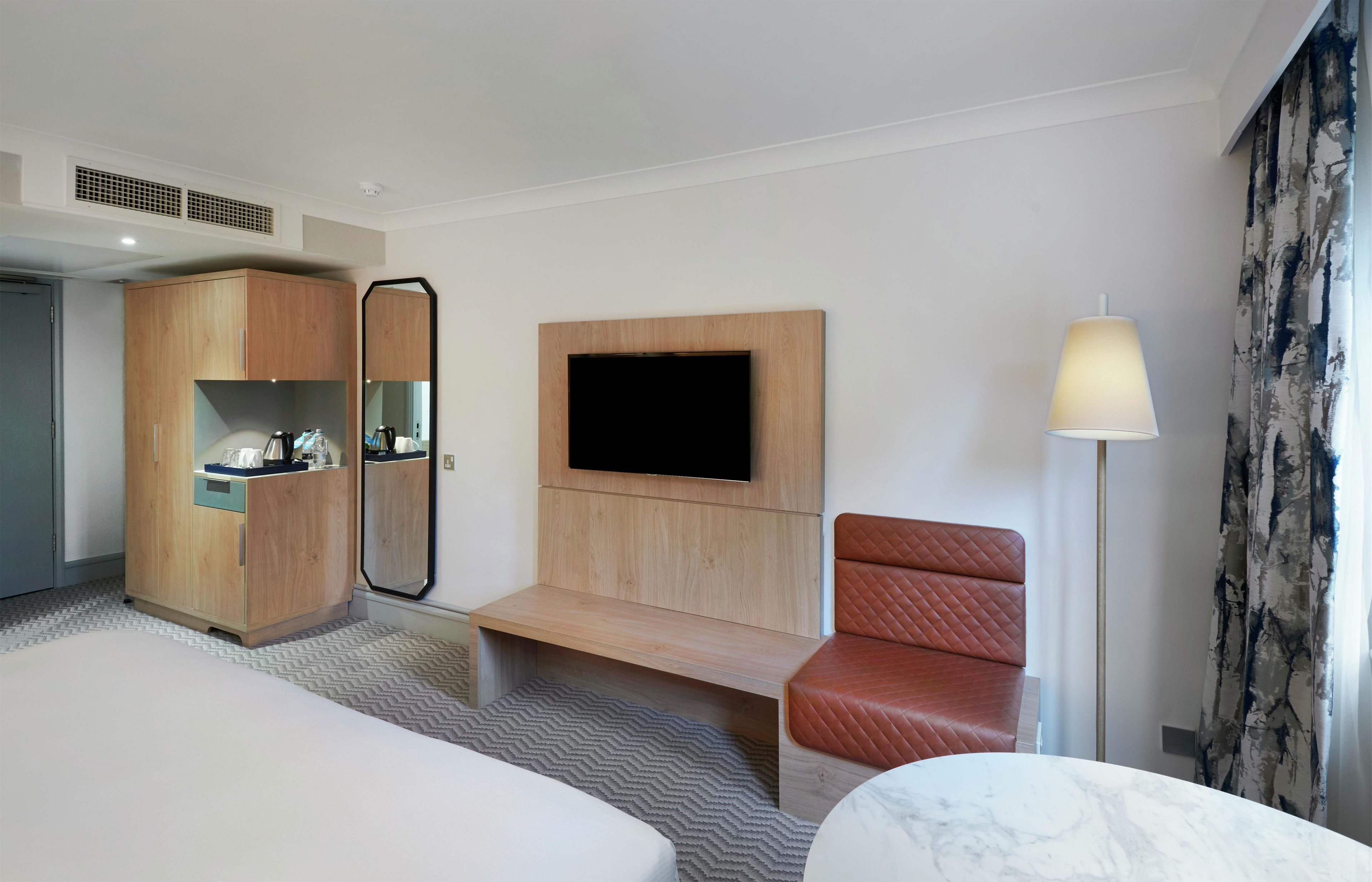 Habitació d'hotel amb un llit, un armari de fusta, una televisió penjada a la paret, una zona de seients amb una cadira i una làmpada.
