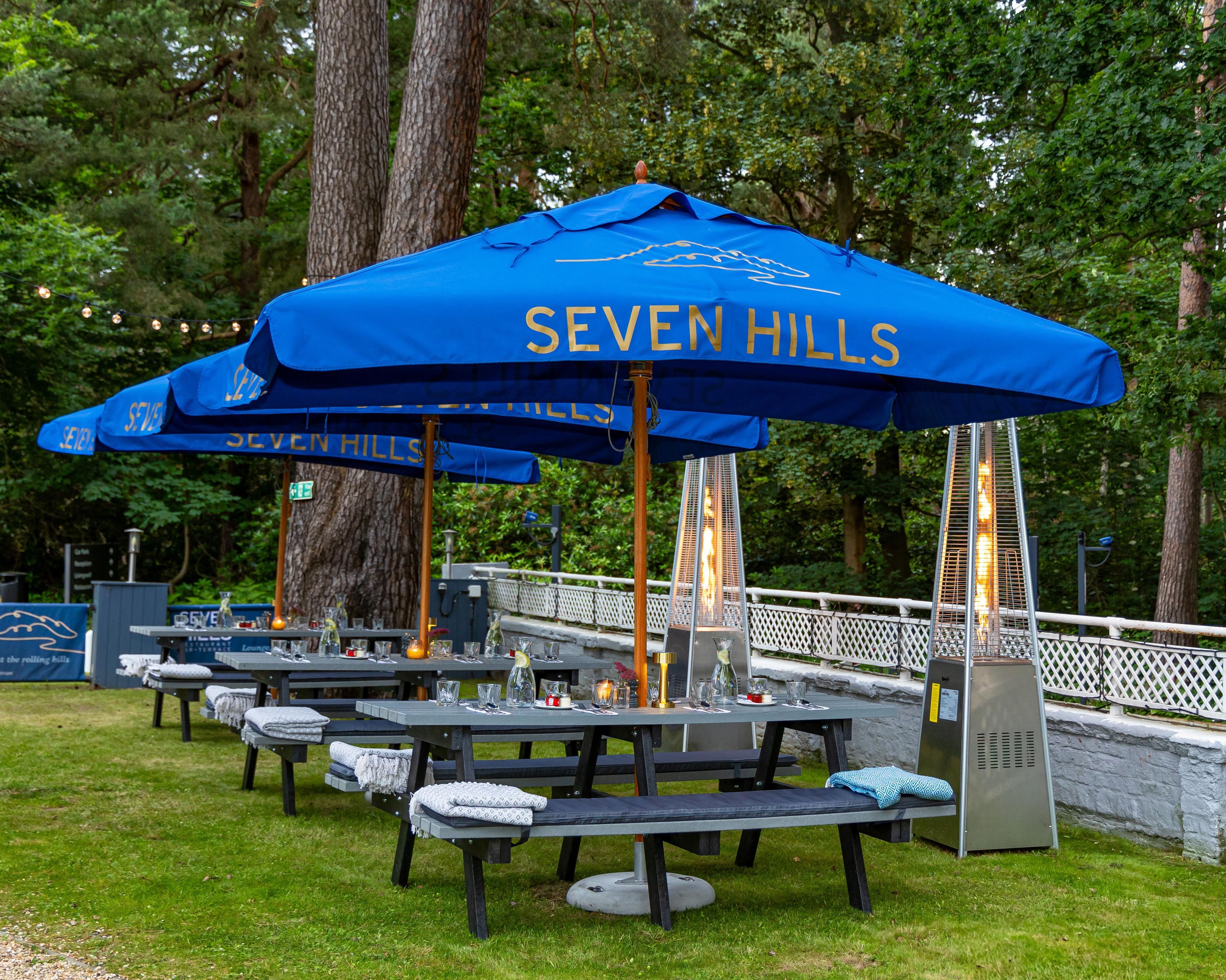 Outdoor seating with blue umbrellas labeled 'SEVEN HILLS' and a metal heater.