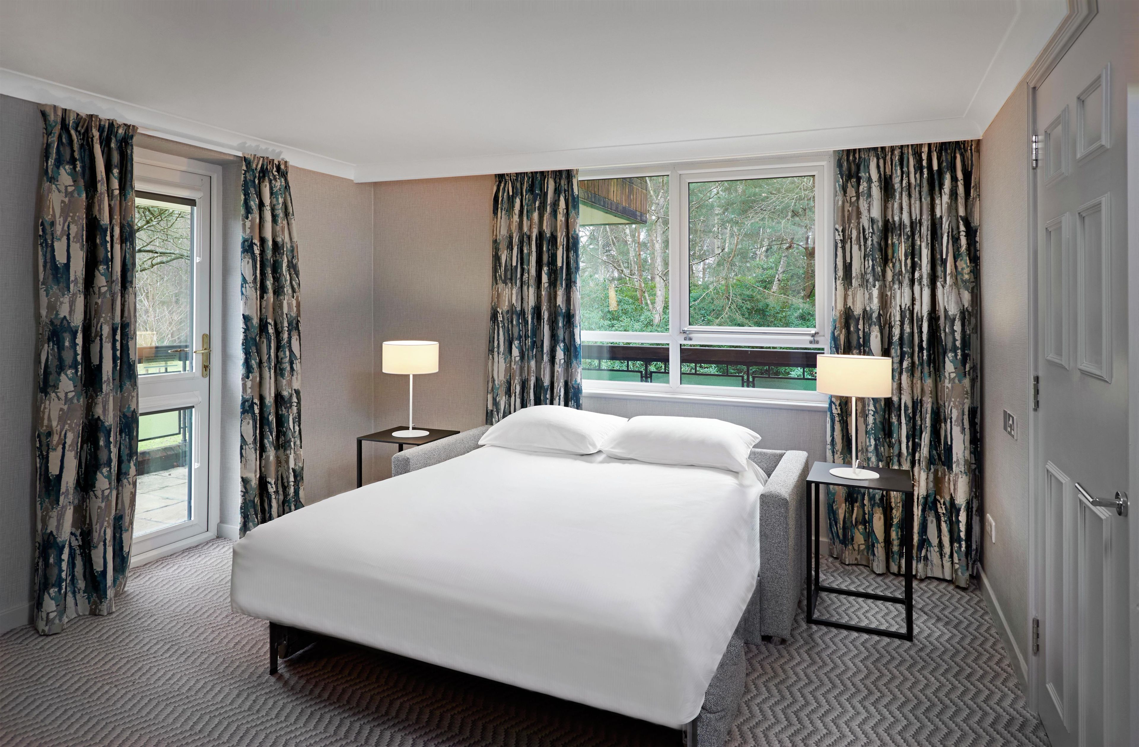Habitación de hotel con cama, dos mesitas de noche con lámparas, ventana con cortinas y vista a zonas verdes.
