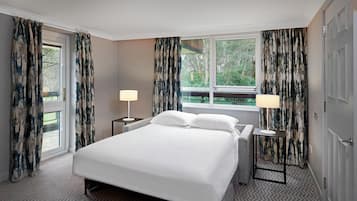 Habitación de hotel con cama, dos mesitas de noche con lámparas, ventana con cortinas y vista a zonas verdes.