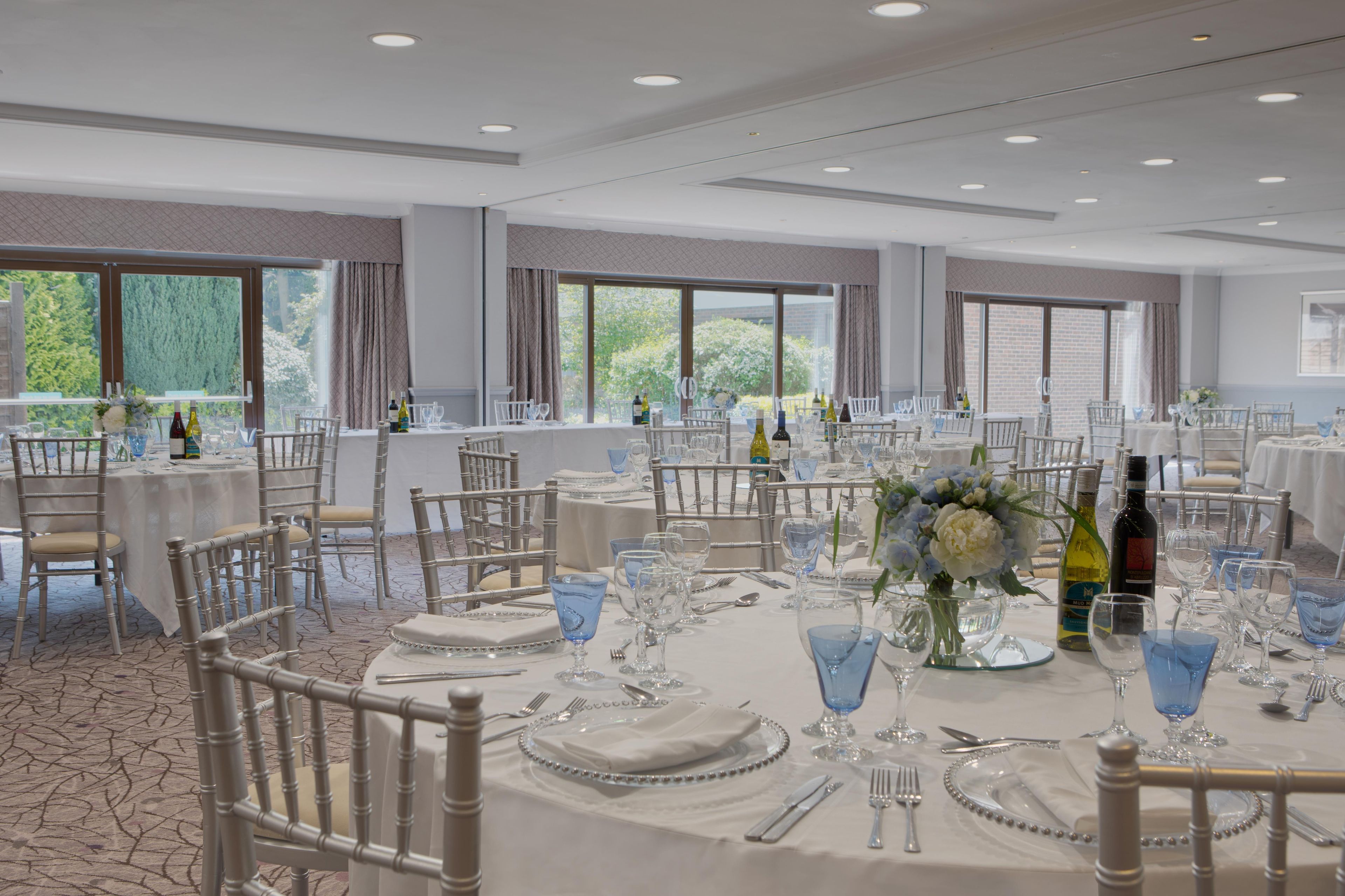 A banquet hall with round tables set for dining, chairs, and a large window providing a view of greenery.