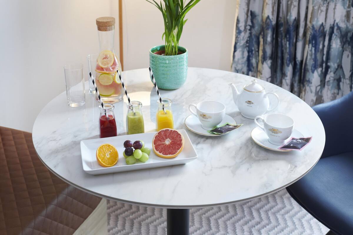 A round marble table with a fruit platter, two glass bottles with straws, a teapot, cups, and saucers.