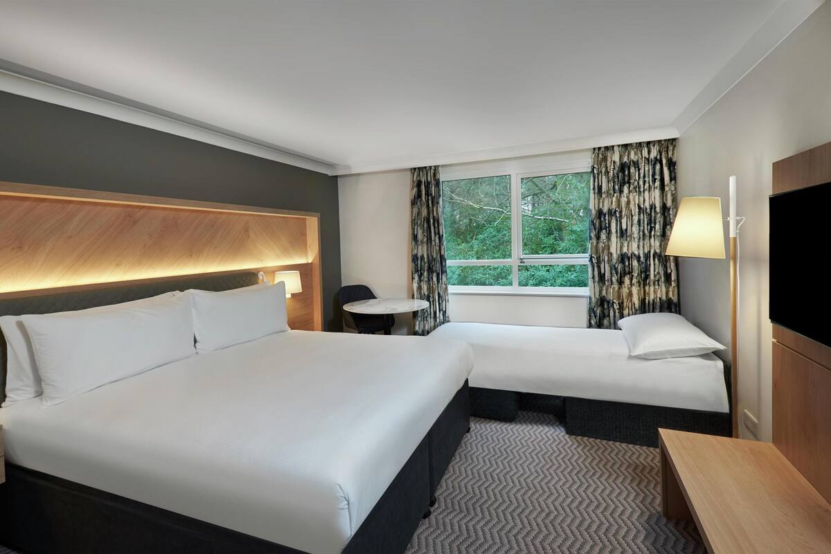 A hotel room with two beds, a small table, a chair, a TV, and a window with curtains.