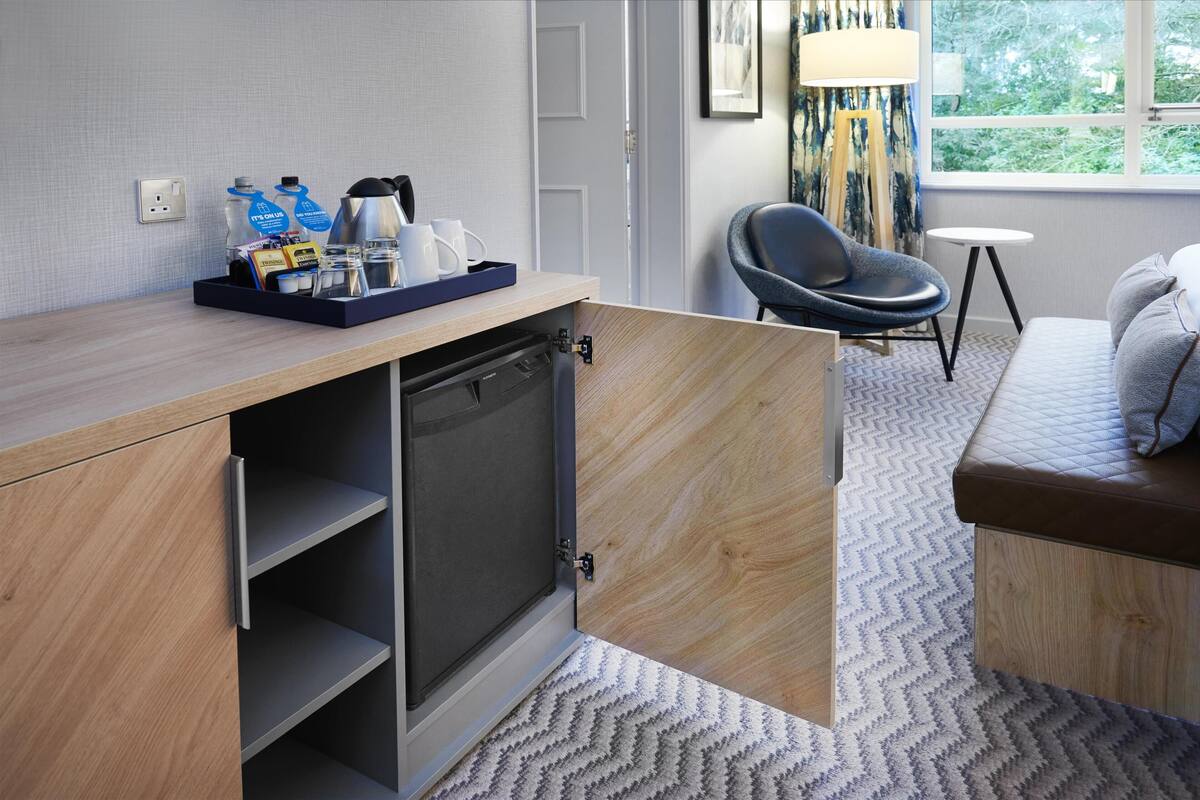 A compact hotel room with a kitchenette, a seating area with a sofa and chair, and a window with a view of greenery.