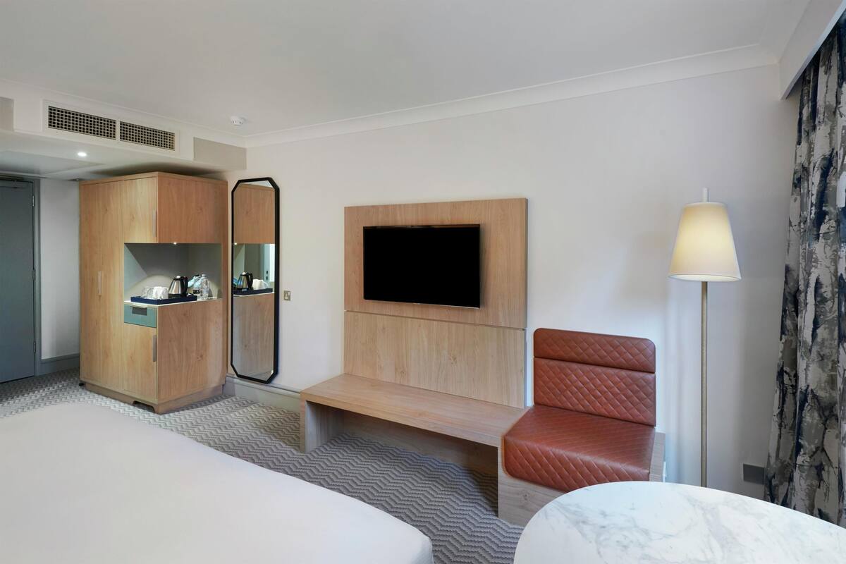 A hotel room with a bed, a wooden wardrobe, a television mounted on the wall, a seating area with a chair, and a lamp.