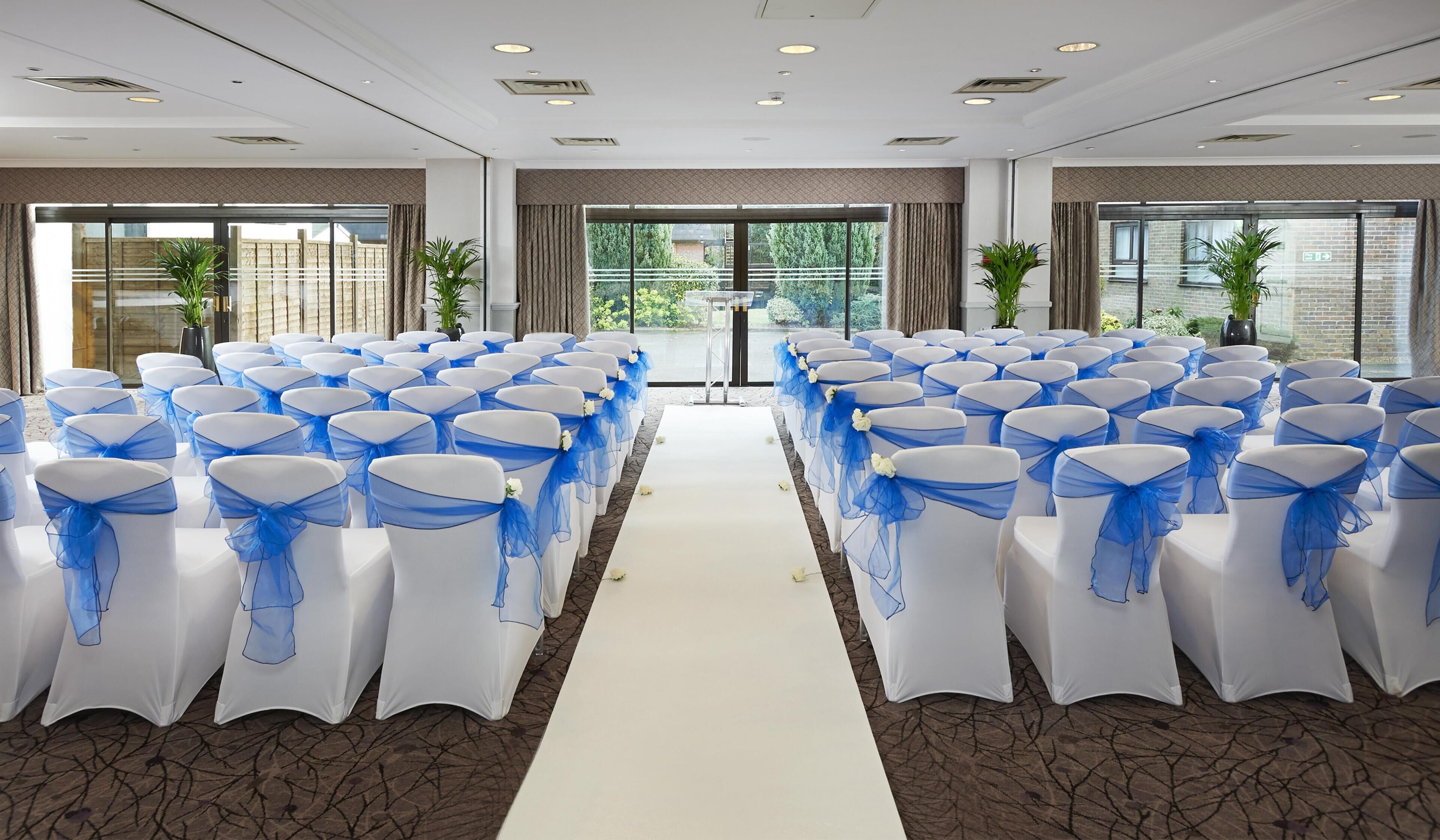 A conference room with rows of white chairs with blue sashes, a white aisle, and large windows with views of greenery.