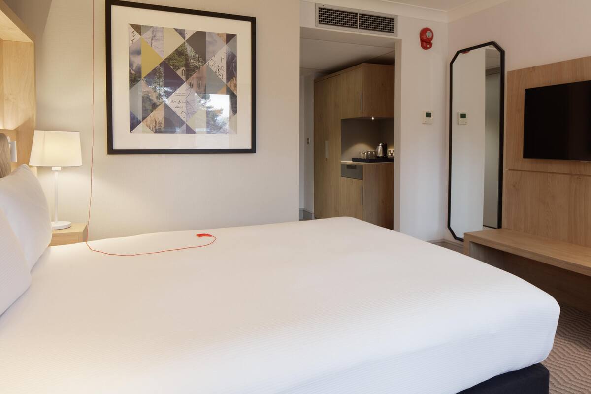 A hotel room with a bed, a bedside lamp, a framed artwork, a mirror, and a television.