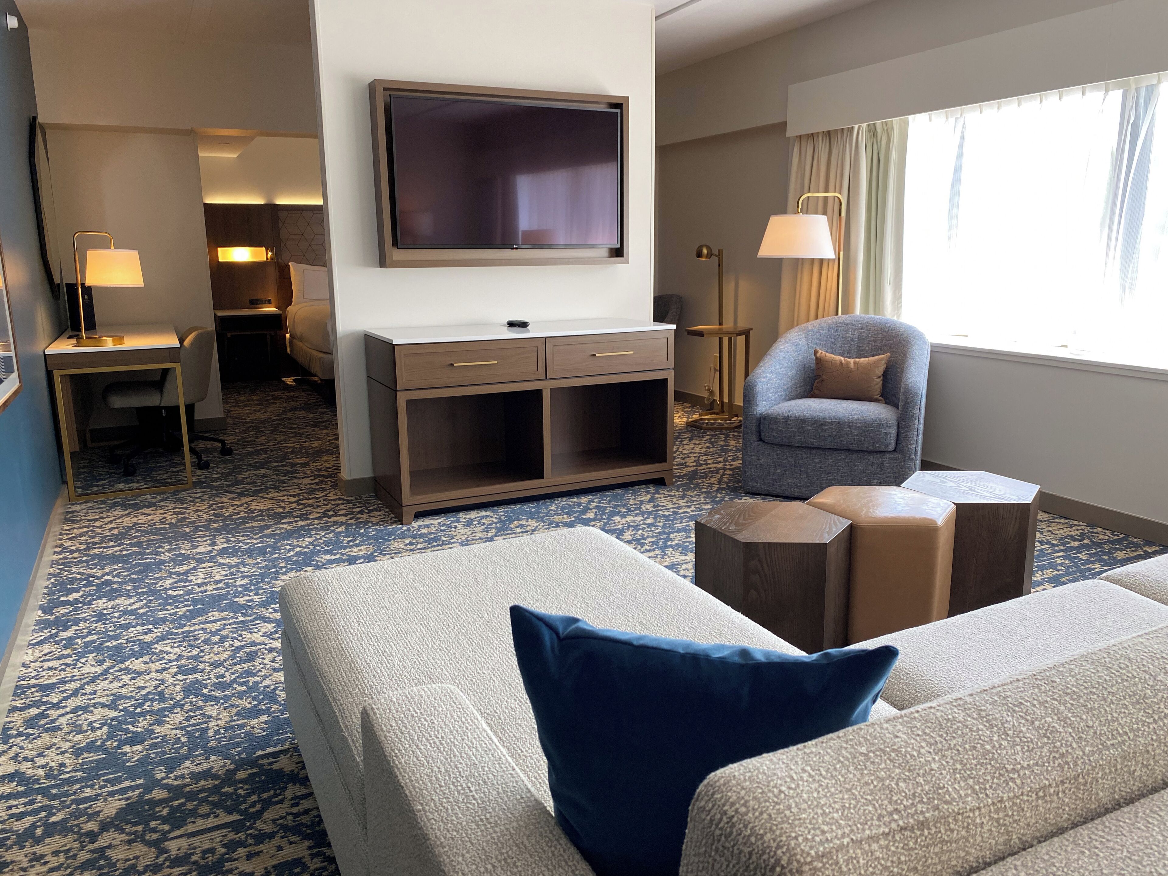 Habitación de hotel con un televisor de pantalla plana grande montado en la pared, un sofá con un cojín azul, una mesita de madera y una silla con cojín.