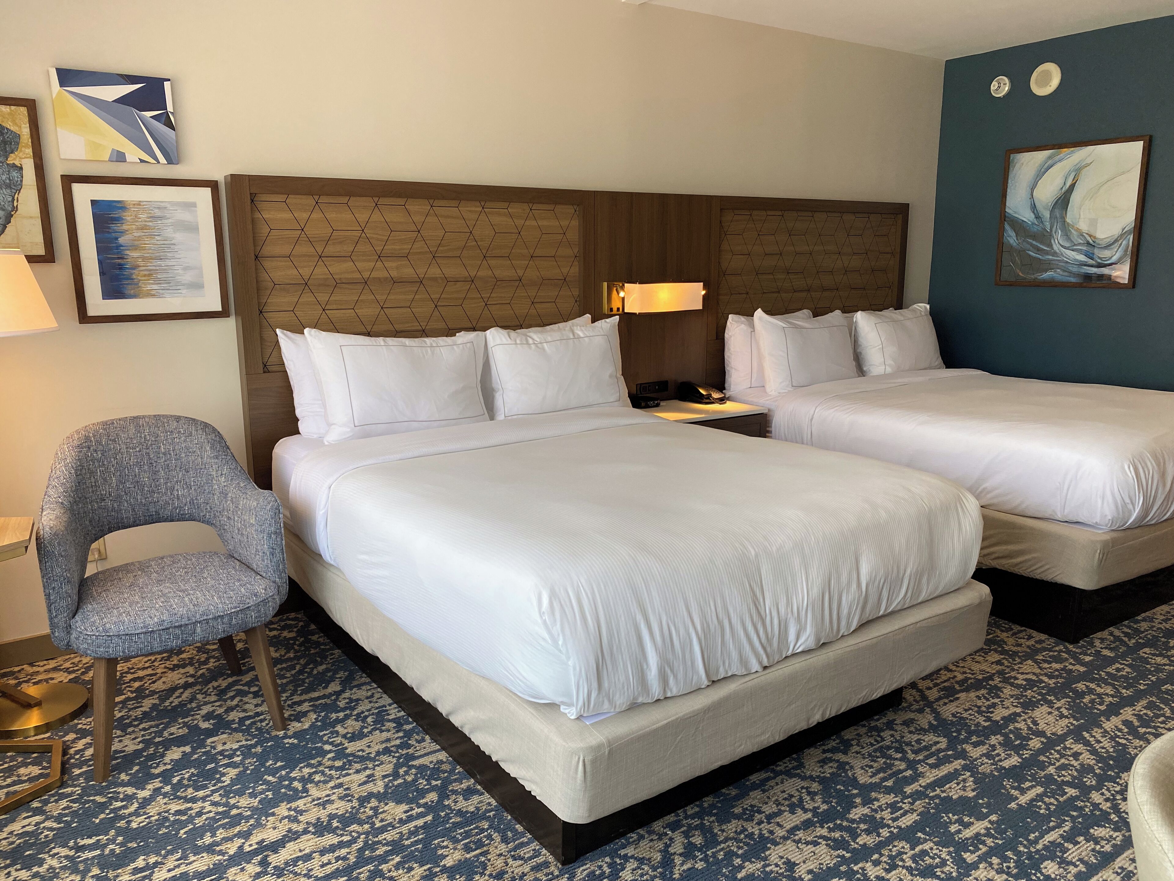 Habitación de hotel con dos camas, una silla, una lámpara y obras de arte en las paredes.