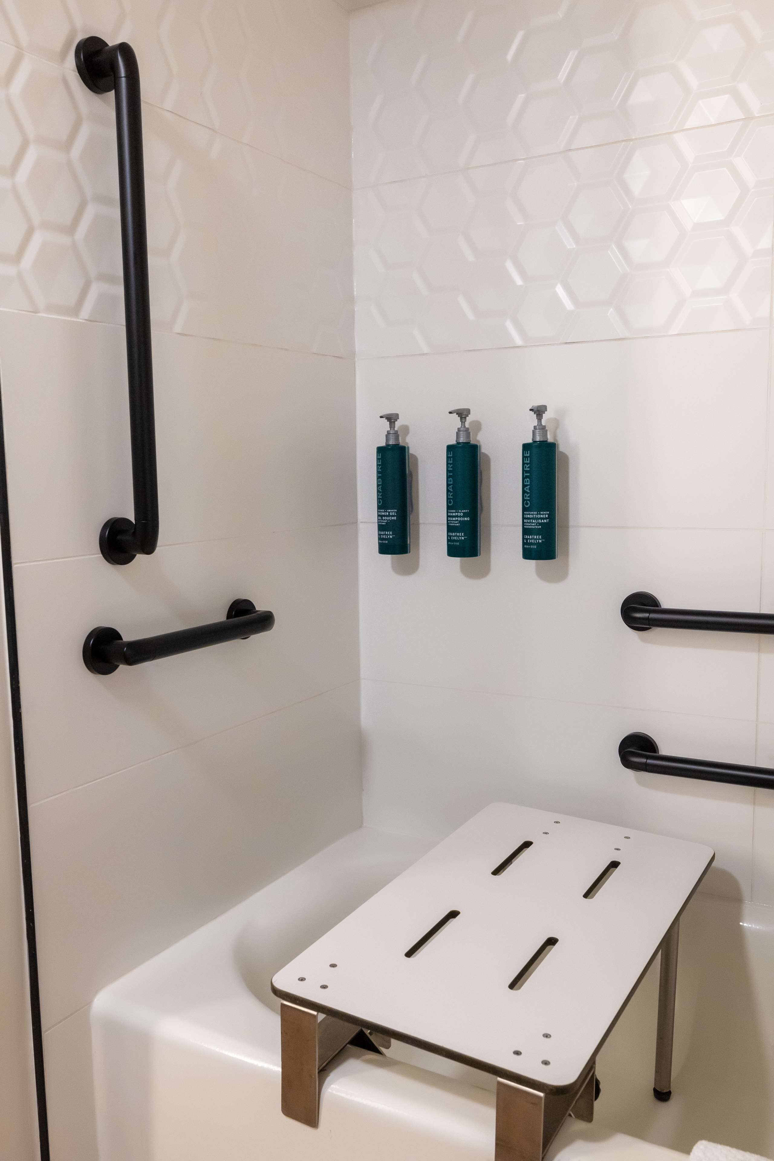A modern bathroom with white tiles, black grab bars, and a white portable shower seat.