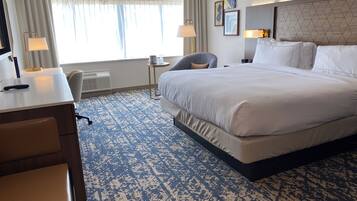 A hotel room with a large bed, a desk with a chair, a small table with a lamp, and a window with curtains.