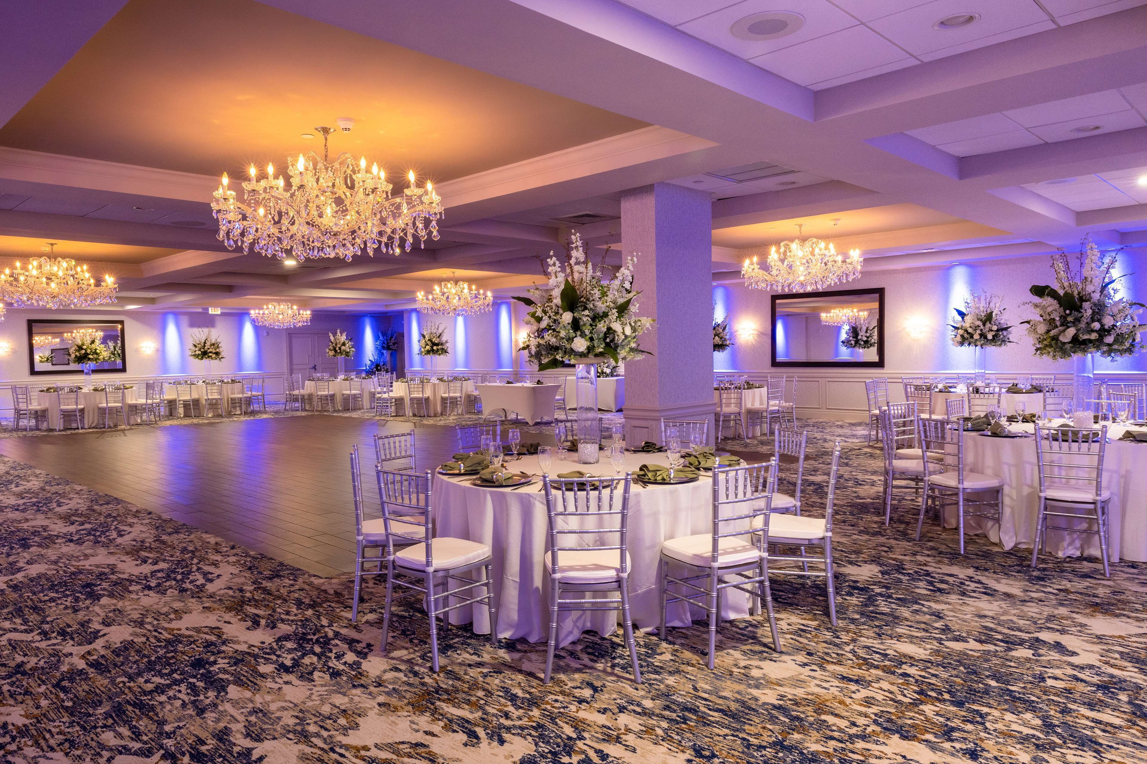A ballroom with round tables, white chairs, chandeliers, and floral centerpieces.