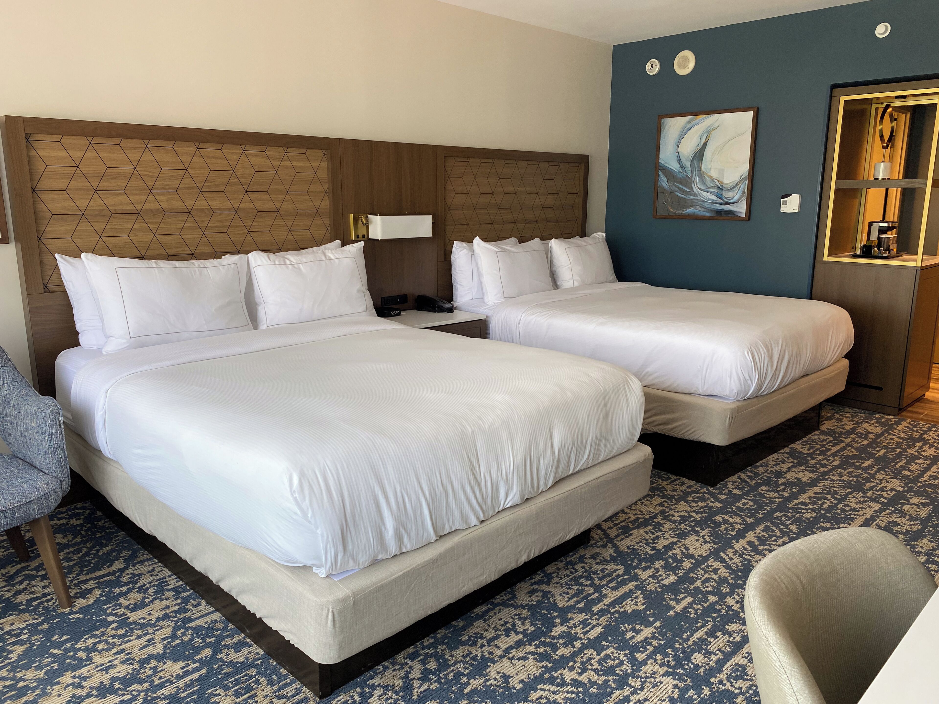 Habitación de hotel con dos camas, cabecero de madera, alfombra azul, un cuadro y un minibar.
