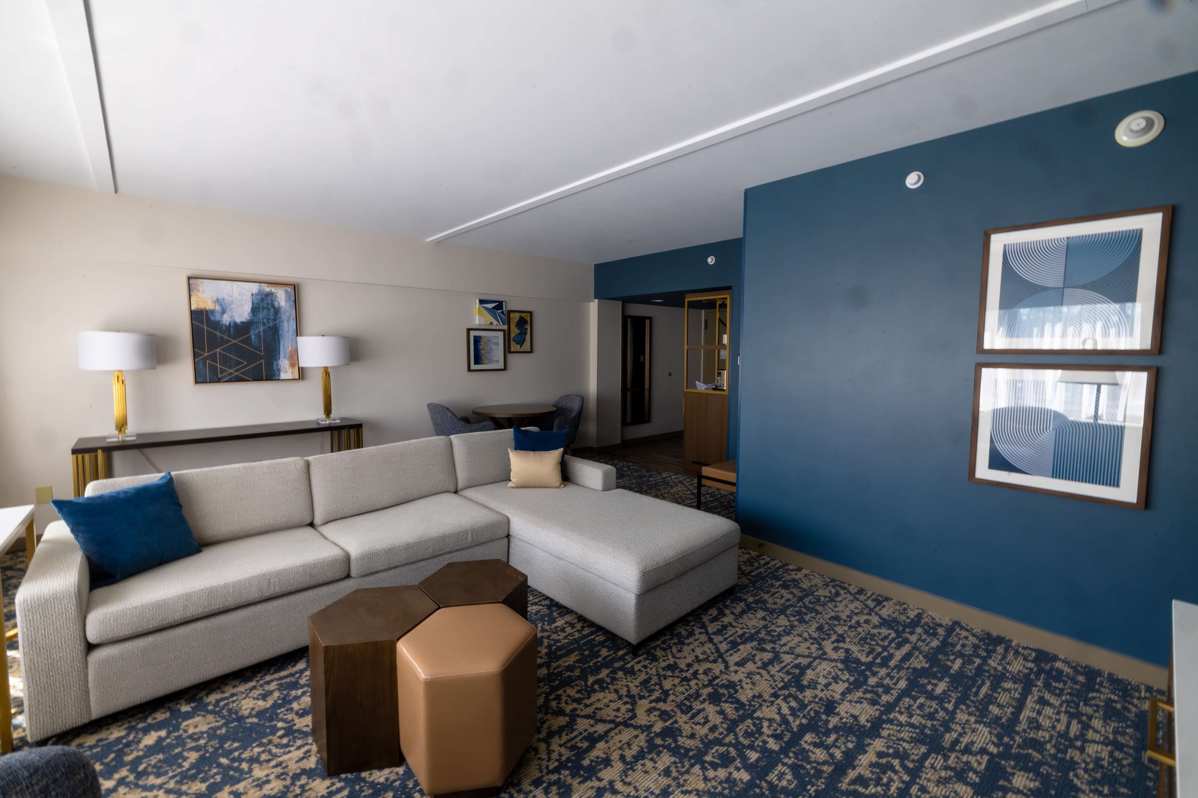 Una habitación de hotel moderna con un sofá seccional gris, un pequeño otoman, una alfombra estampada azul y obras de arte en las paredes.