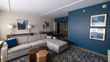Una habitación de hotel moderna con un sofá seccional gris, un pequeño otoman, una alfombra estampada azul y obras de arte en las paredes.