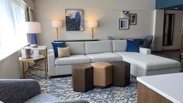 A modern hotel room with a grey sectional sofa, two side chairs, a wooden coffee table, and a patterned carpet.