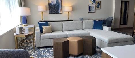 A modern hotel room with a grey sectional sofa, two side chairs, a wooden coffee table, and a patterned carpet.